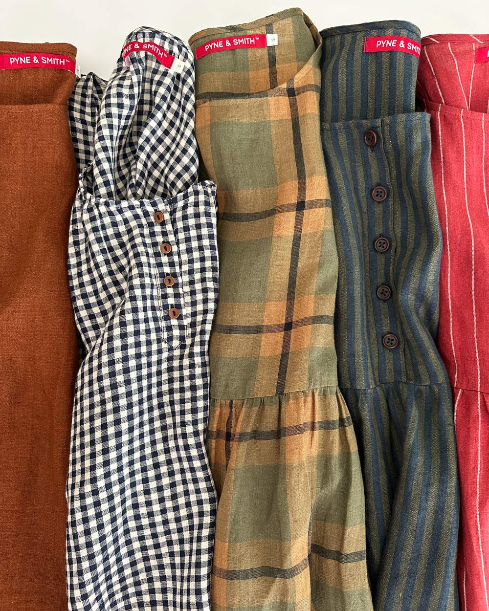 Five Pyne & Smith dresses are laid side by side, each featuring different fabric patterns: solid rust, black and white gingham, tan and green plaid, green with thin dark stripes and buttons, and red with white pinstripes.