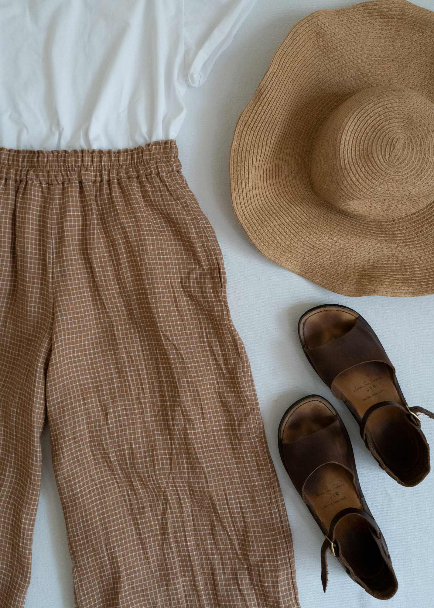 A flat lay of a white short-sleeve top, brown plaid pants, a wide-brimmed straw hat, and a pair of brown leather sandals on a light surface.
