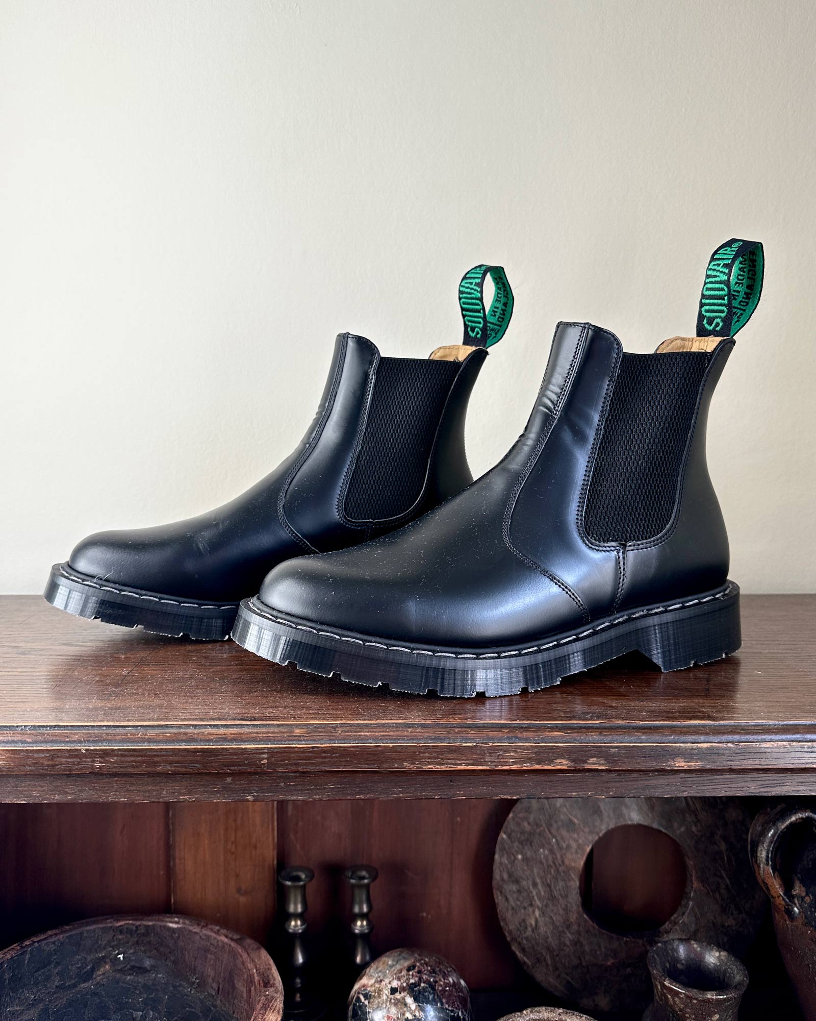 A pair of Solovair Black Leather Dealer Boots by Pyne and Smith Clothiers, with chunky Goodyear welted soles and green pull tabs, rest on a wooden surface against a light-colored wall.