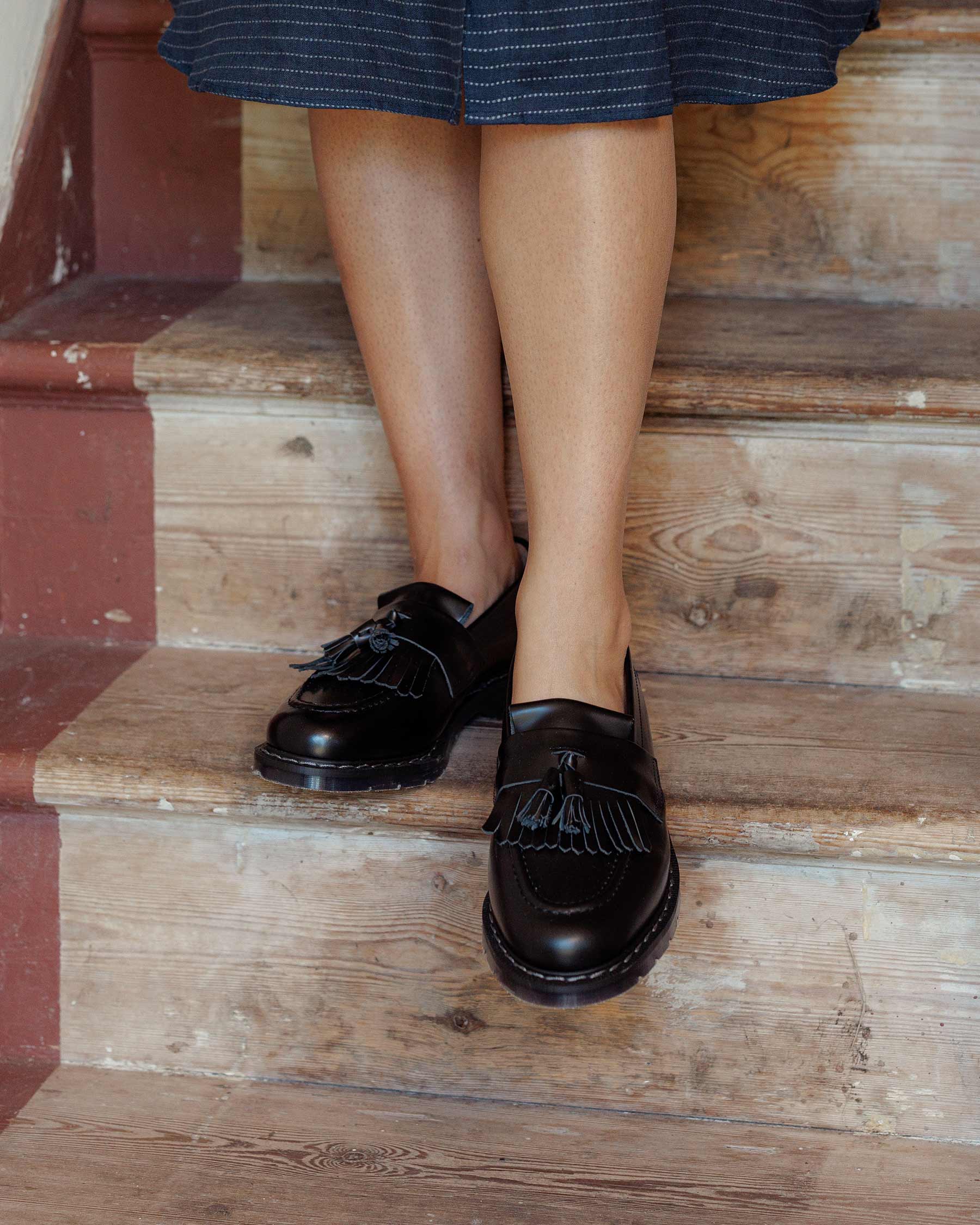 A person wearing a dark skirt and Pyne and Smith Clothiers Solovair Black Leather Hi-Shine Tassle Loafers stands on worn wooden stairs.