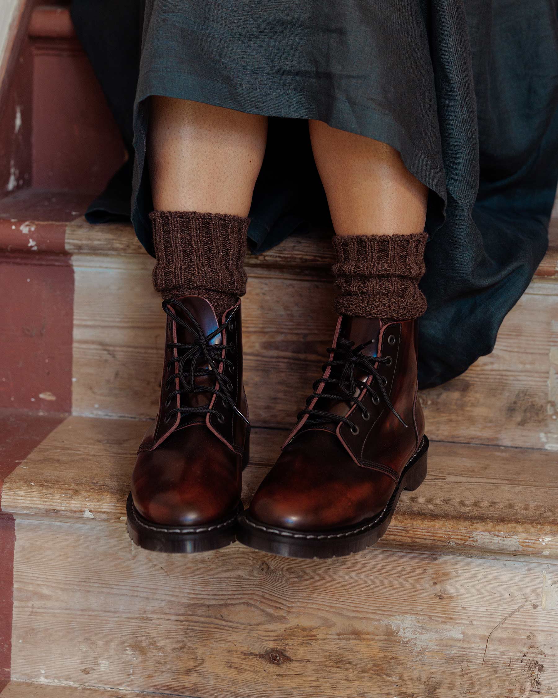 A person wearing Pyne and Smith Clothiers’ Solovair Burgundy Leather Astronaut Boot with a Goodyear welted sole and brown knitted socks sits on wooden stairs, dressed in a dark skirt.