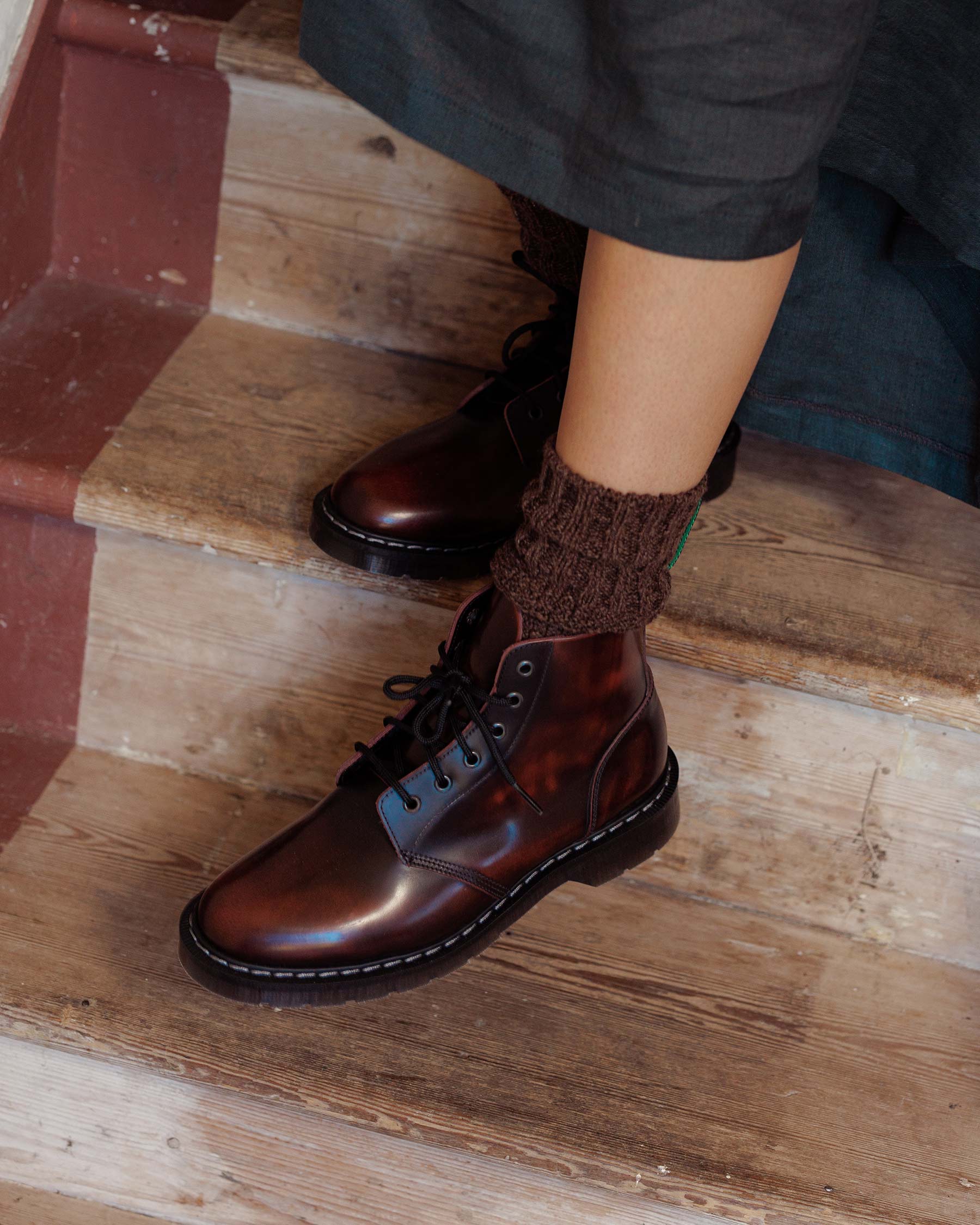 A person wearing Pyne and Smith Clothiers Solovair Burgundy Leather Astronaut Boots with brown knitted socks sits on a worn wooden staircase, one foot on a step and part of a dark skirt visible.