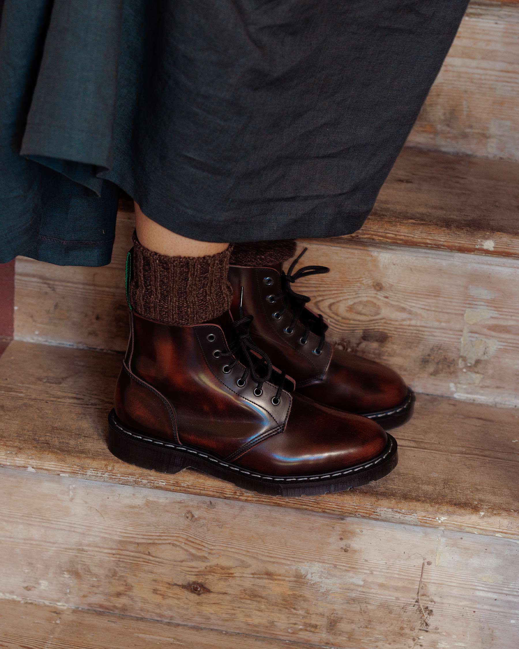 A person wearing Pyne and Smith Clothiers Solovair Burgundy Leather Astronaut Boot with brown socks stands on wooden stairs, part of a dark skirt or dress visible. The boots have a Goodyear welted sole for durability.