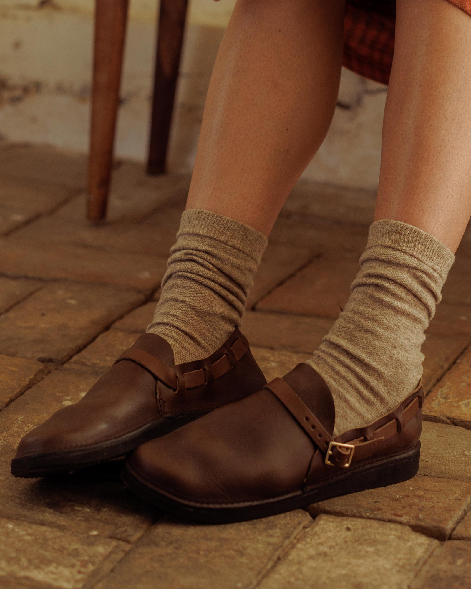 A person wearing Pyne and Smith Clothiers Cashmere Wool Socks in Beige and brown leather buckle shoes sits on a tiled floor. Only their lower legs and feet are visible.