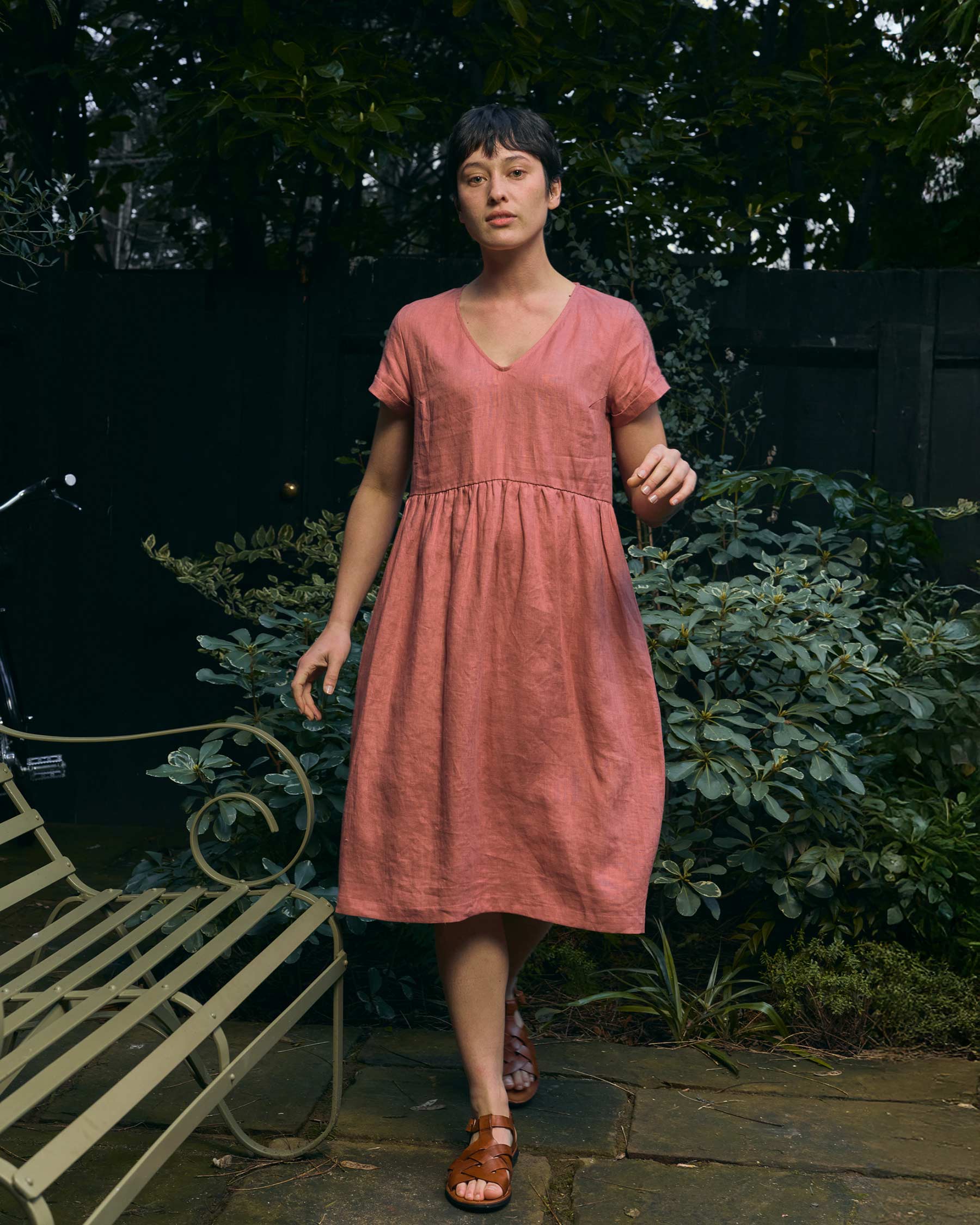 A woman with short dark hair walks in a leafy garden past a metal bench, wearing Pyne and Smith Clothiers No.40 V Neckline Linen Day Dress in Brick Dust and brown sandals.