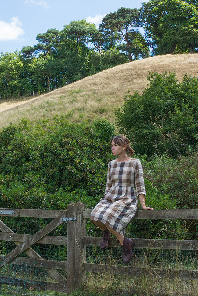 A woman in a plaid dress sits on a wooden fence in front of grassy hills and green trees under a partly cloudy sky.