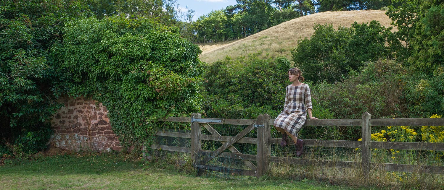 A person in a plaid dress sits on a wooden gate surrounded by lush green vegetation, with a stone wall to the left and a grassy hill in the background under a partly cloudy sky.