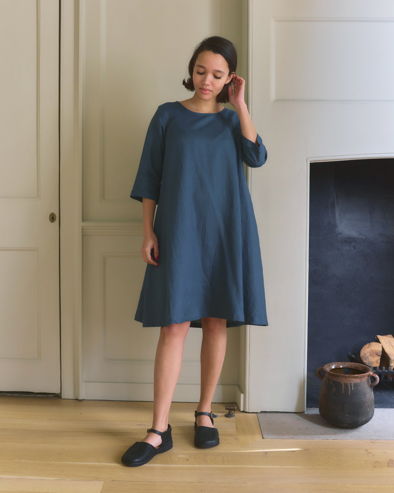 A woman with short dark hair stands indoors on a wooden floor, touching her hair and looking down. She wears the No.29 A-line Linen Dress in Stargazer by Pyne and Smith Clothiers, with black shoes, near a fireplace with logs and a brown pot.
