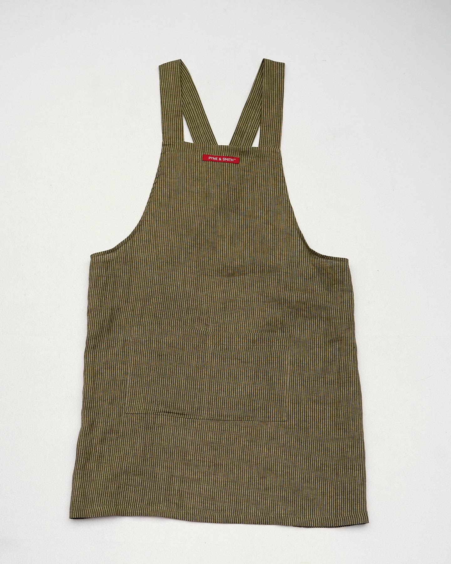 The Cross Back Apron in Olive Stripe Linen by Pyne and Smith Clothiers features wide straps, a brown and green pinstripe design, ethically made European linen, and a red top label, shown laid flat on a white background.