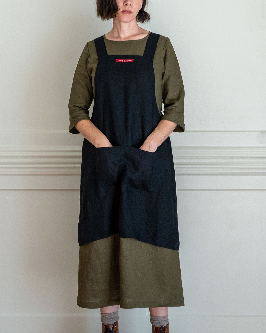 A woman stands against a light wall, wearing an olive green dress and the Pyne and Smith Clothiers Cross Back Apron in Black Linen, featuring large front pockets and a red and white chest label. Her hands are in the pockets.