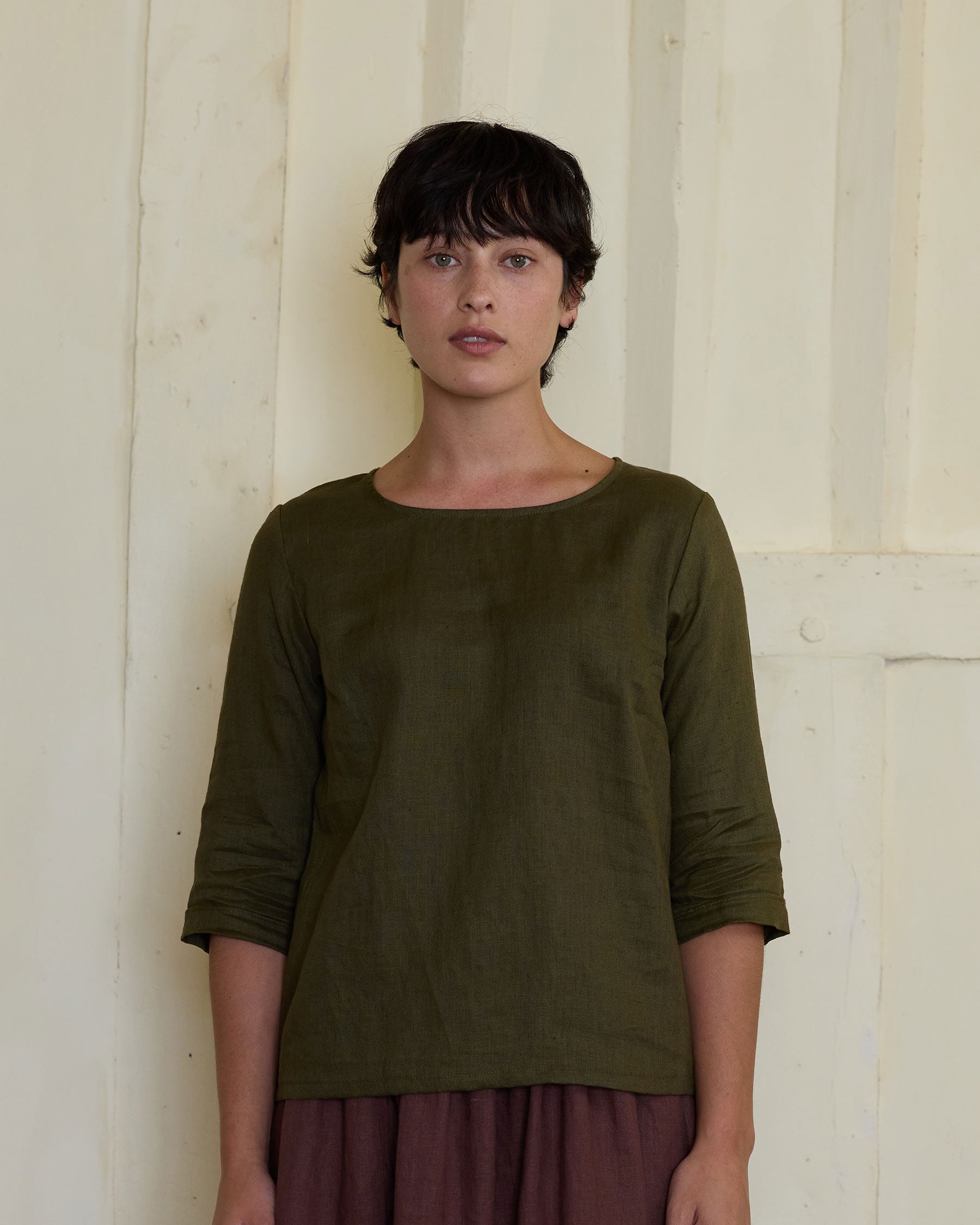 A person with short dark hair stands facing forward, wearing the Pyne and Smith Clothiers Blouse No.12 Everyday Linen Top in Beech and a reddish-brown skirt, in front of a cream-colored wooden wall.