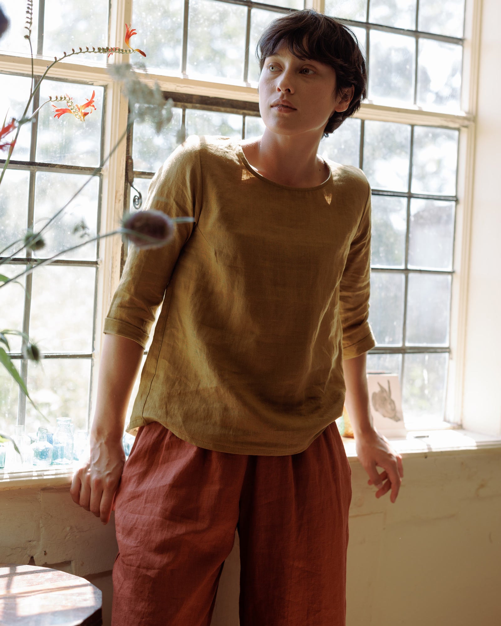 A person with short dark hair stands by a sunlit window, wearing the Pyne and Smith Clothiers Size XS #622 Blouse No.12 Everyday Linen Shirt in Cumin and rust-colored pants. Plants and glass bottles sit on the windowsill nearby.