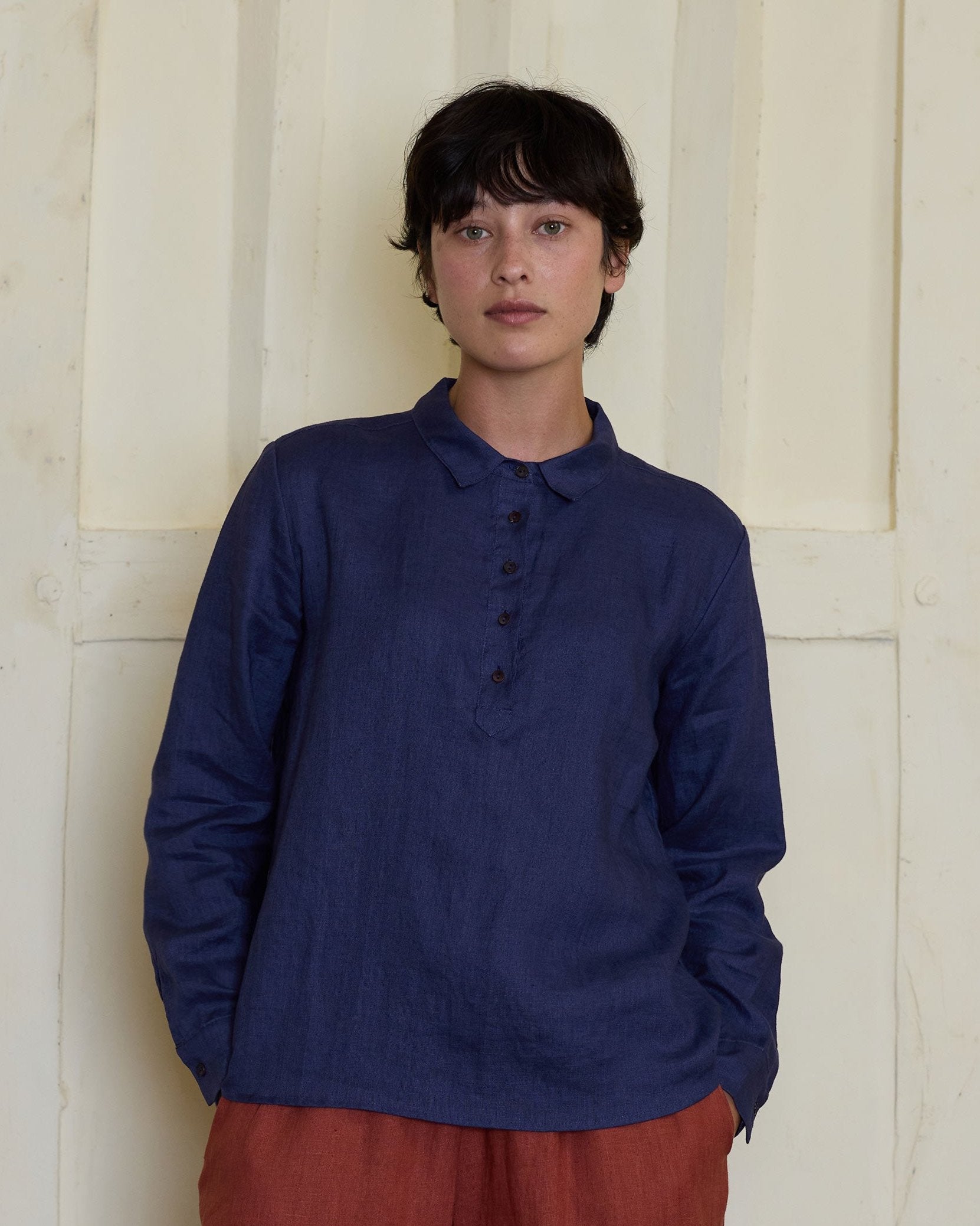 A person with short dark hair wears the Pyne and Smith Clothiers Linen Blouse No.18 Collared Long Sleeved Shirt in Medieval Blue, paired with loose rust-colored pants, standing against a cream textured wall, evoking classic British style.