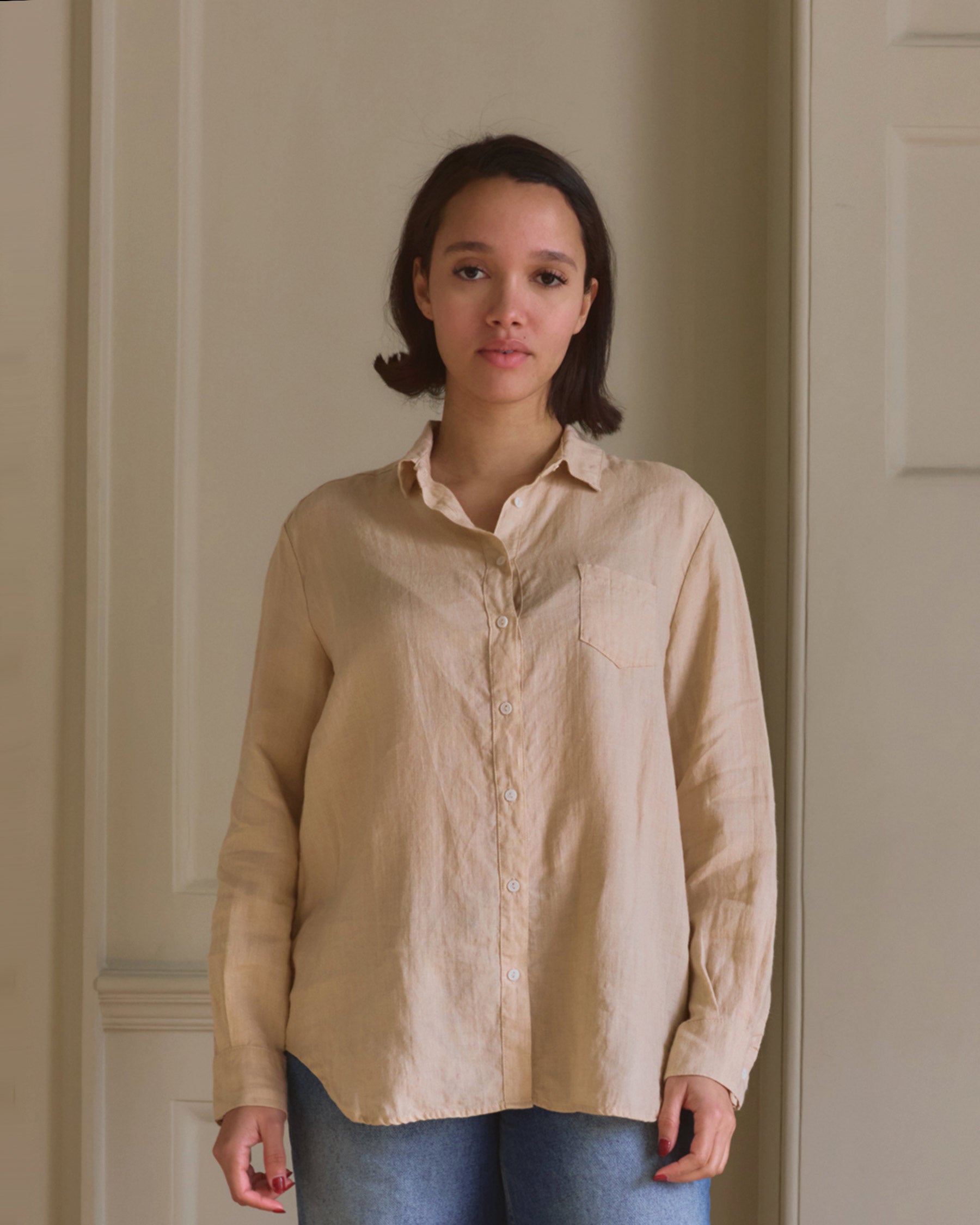 A person with short dark hair stands indoors against a beige wall, wearing the Pyne and Smith Clothiers Shirt No.21 Button up, Linen Blouse in Biscuit with blue jeans, looking directly at the camera with a neutral expression.