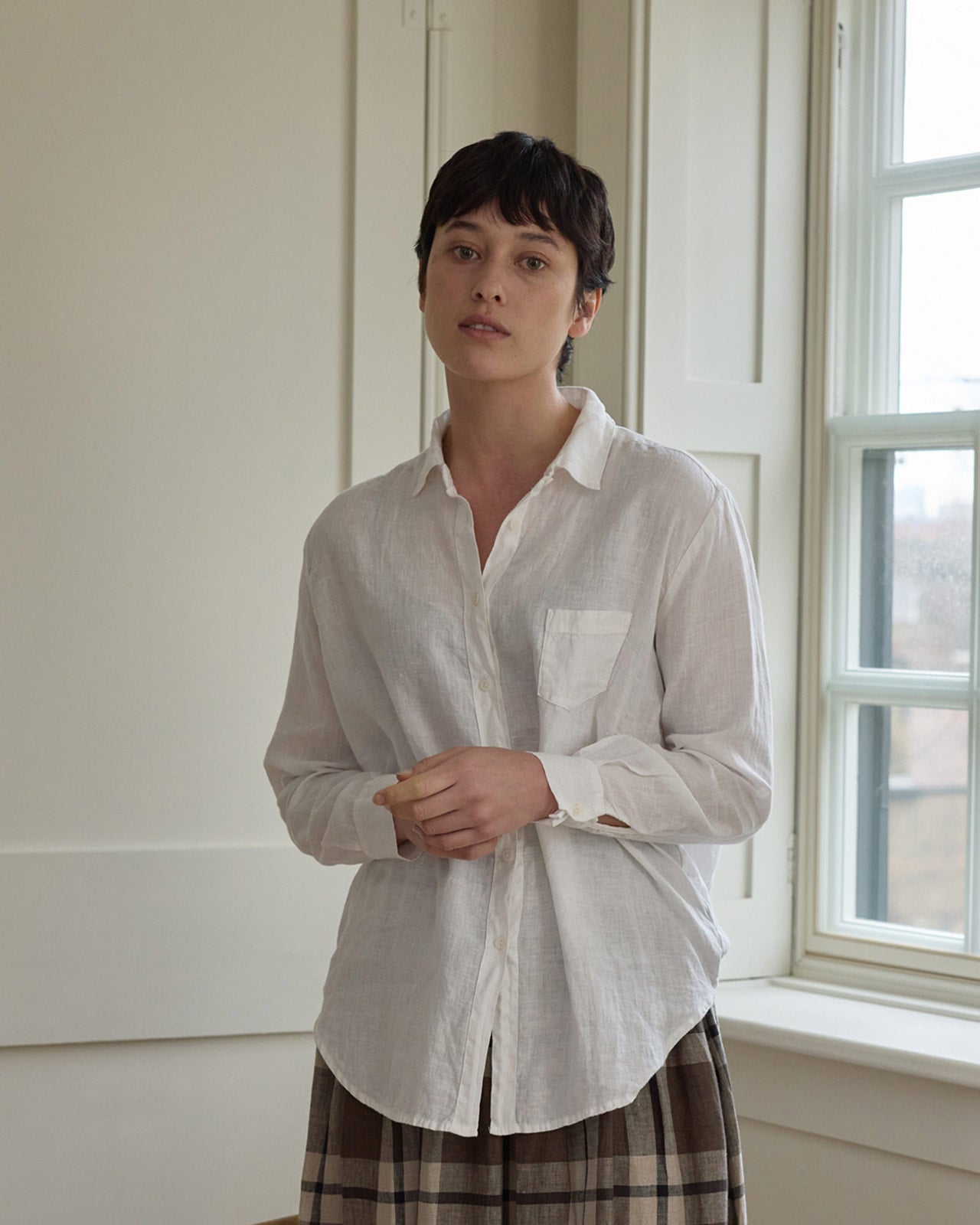 A person with short dark hair wears the Pyne and Smith Clothiers Shirt No.21 Button Up, a linen blouse in soft white, paired with a brown plaid skirt, standing indoors by a sunlit window with neutral white walls and shutters.