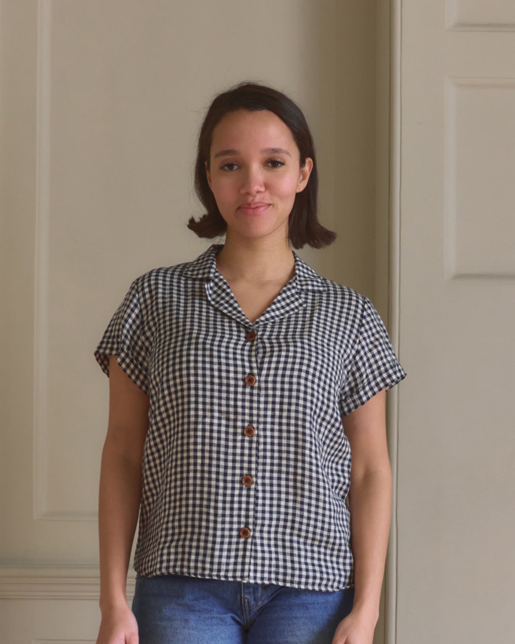 A woman with short dark hair stands indoors against a light wall, wearing Pyne and Smith Clothiers V-Neck Linen Blouse No.19 in Navy Gingham styled as a button-up shirt with blue jeans, looking at the camera.