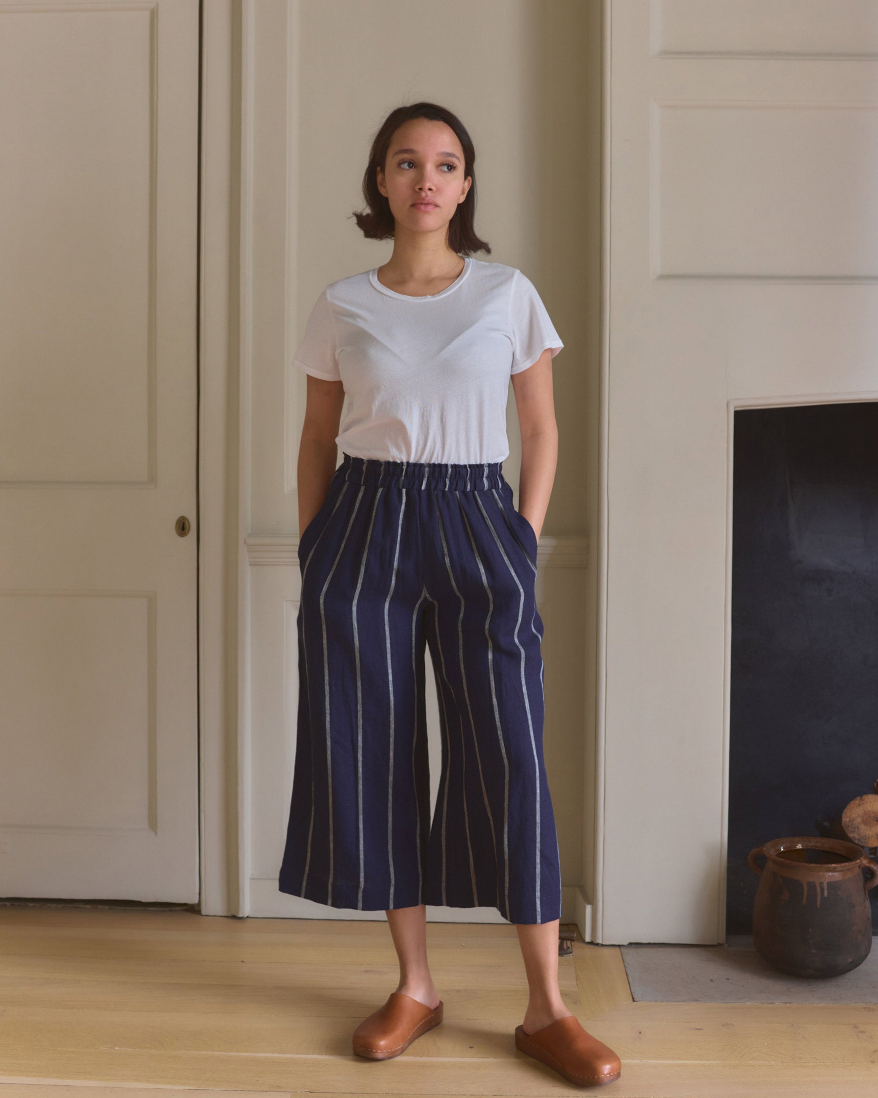 A woman stands indoors in a white tee, brown slip-ons, and Pyne and Smith Clothiers Culottes No.11 Linen Cropped Pants in Blundell Stripe, hands in her pockets before a white paneled wall and fireplace.