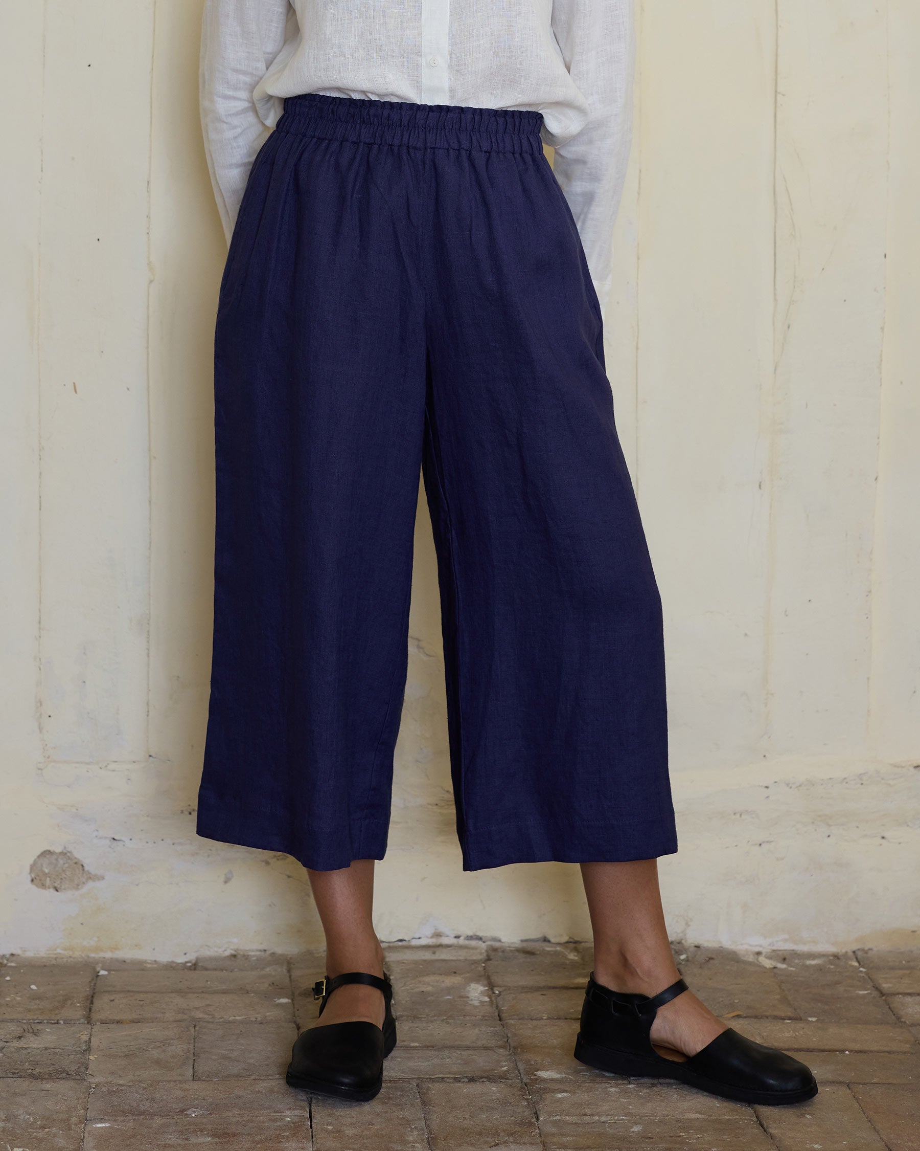 A person stands against a light wall, wearing a white blouse, black Mary Jane shoes, and Pyne and Smith Clothiers Culottes No.11 Linen Cropped Pants in Medieval Blue with an elasticated waistband. Their hands are in their pockets, head out of frame.