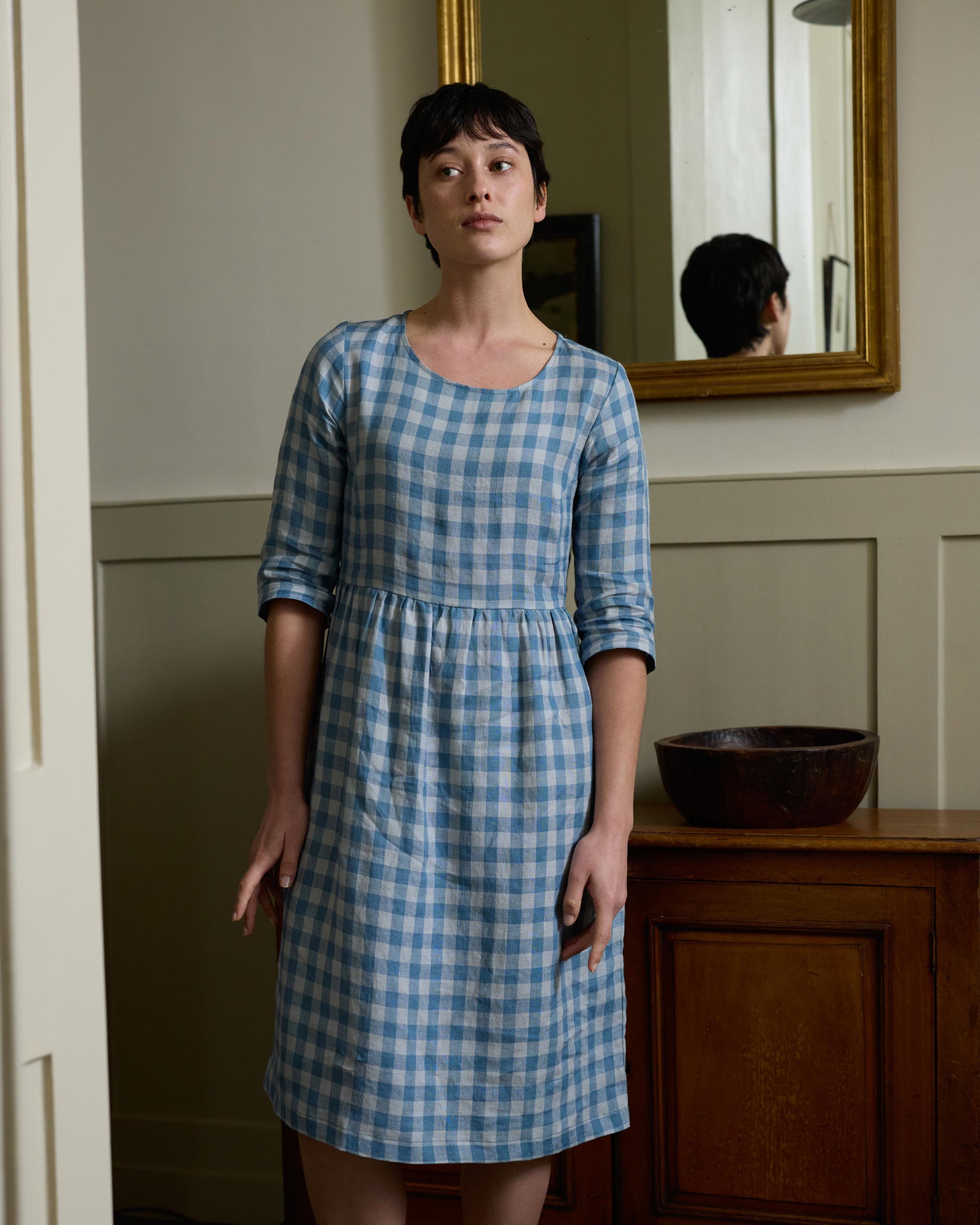 A person with short dark hair stands indoors near a wooden cabinet and mirror, wearing the Pyne and Smith Clothiers No.19 Boatneck Midi Linen Dress in Bothy Check, which features an adjustable waist tie, looking slightly to the side.