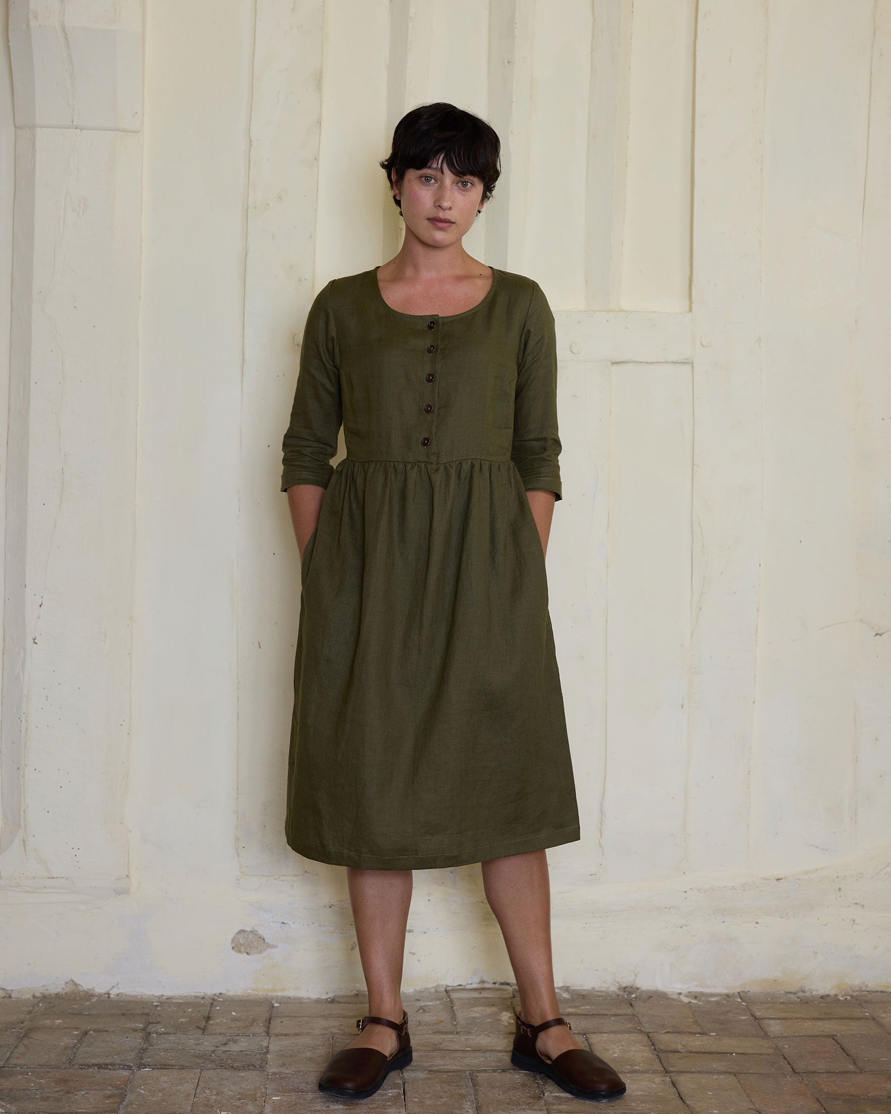 A person with short dark hair stands against a textured cream wall, wearing the No.12 Button Up Linen Midi Dress in Beech by Pyne and Smith Clothiers—a sustainable, effortless look—with hands in pockets and looking directly at the camera.