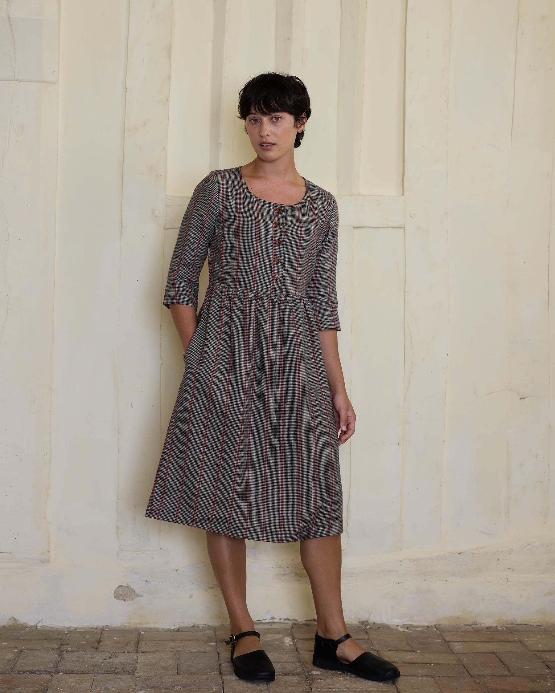 A woman with short dark hair wears the Pyne and Smith Clothiers No.12 Button Up Linen Midi Dress in Kings Cross—knee-length, grey with red stripes and front buttons—styled with black Mary Jane shoes against a pale textured wall.