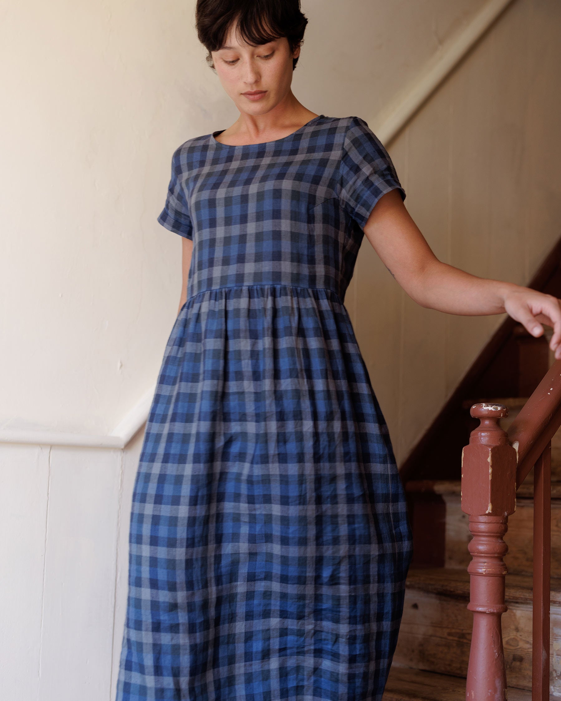 A woman wearing the Pyne and Smith Clothiers No.17 Full Length Linen Tea Dress in Blue Melange stands by a wooden staircase indoors, resting one hand on the railing and looking down in soft lighting.