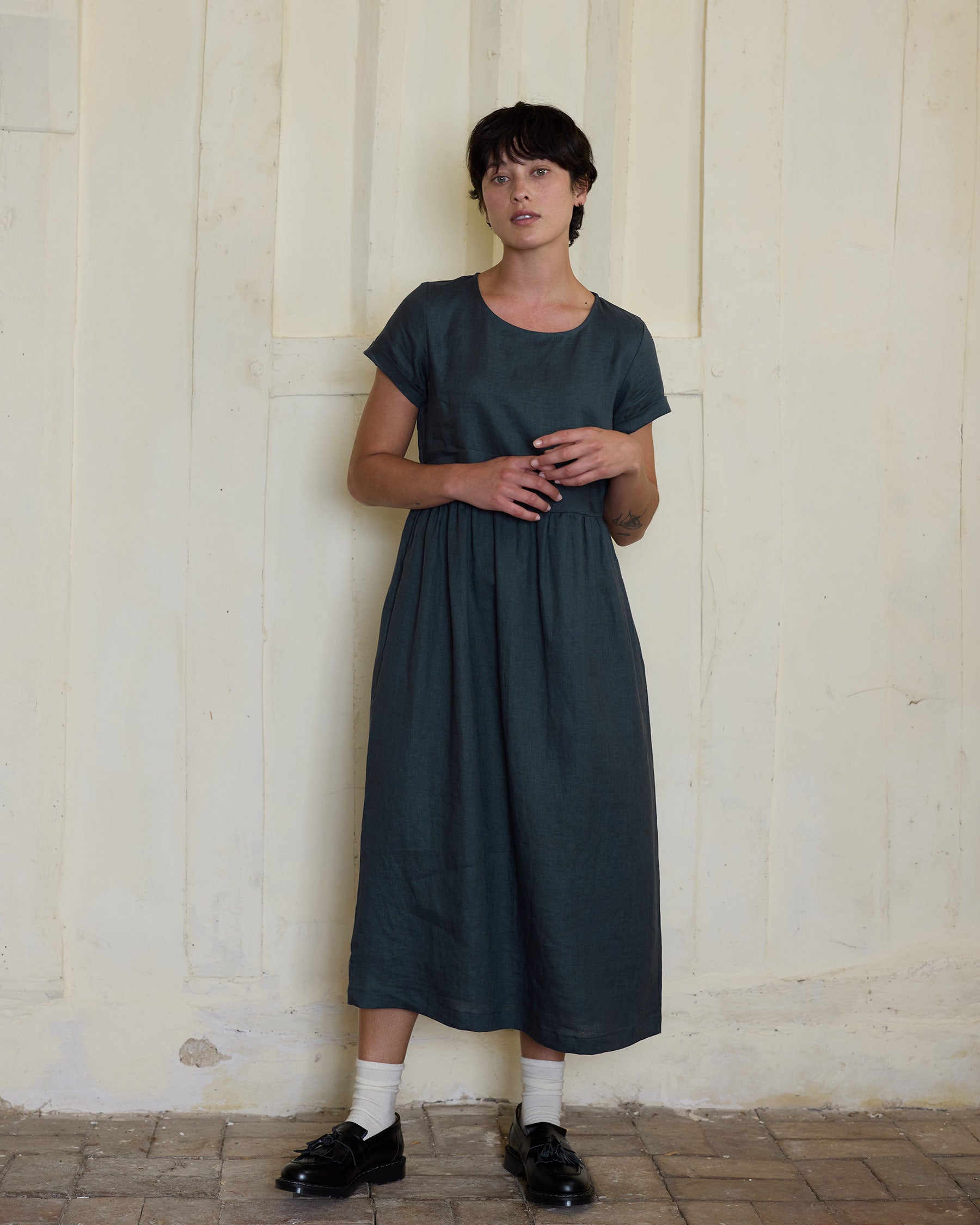 A person with short dark hair stands indoors by a cream textured wall, wearing Pyne and Smith Clothiers No.17 Full Length Linen Tea Dress in Green Gables, styled with black shoes and white socks.