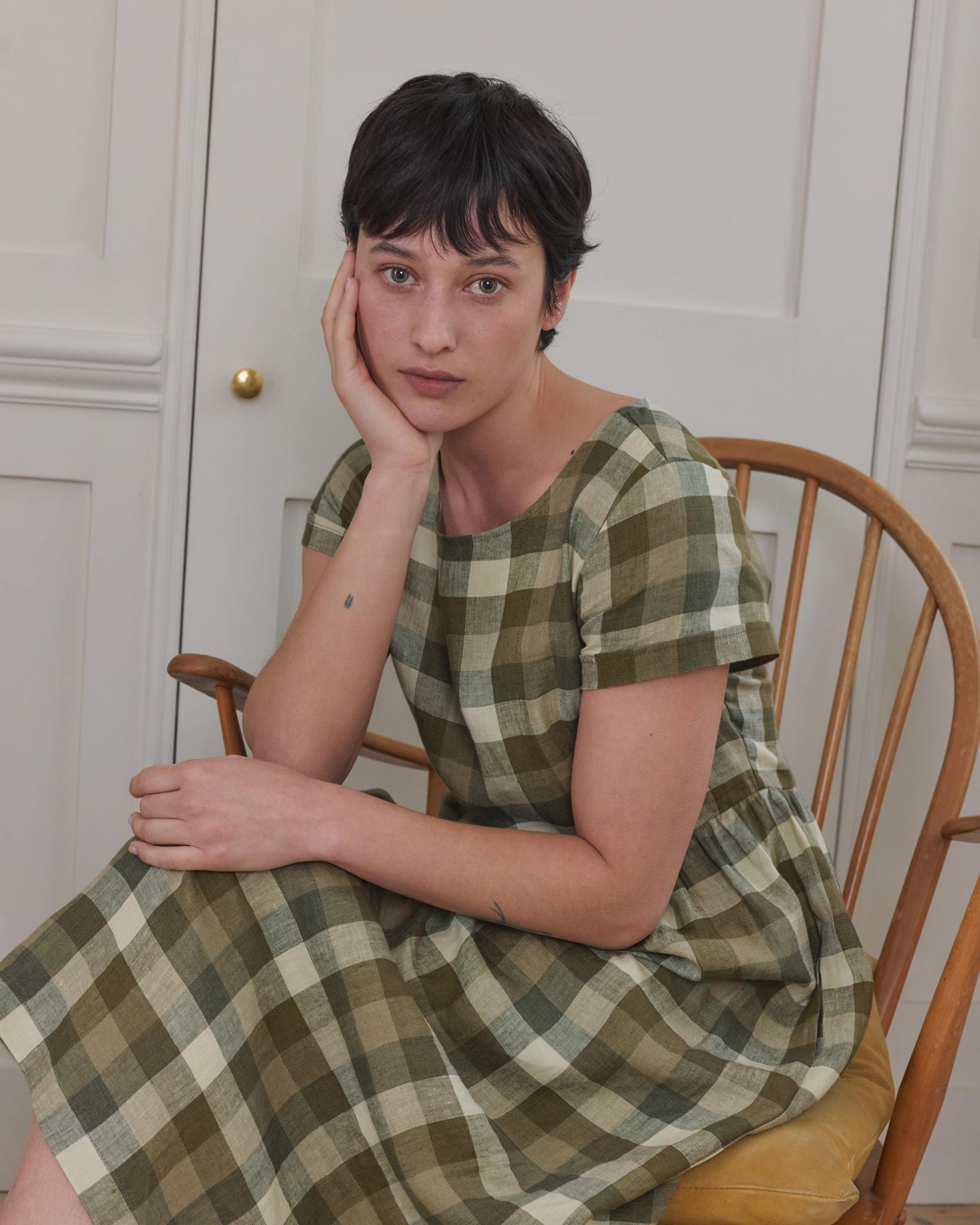 A person with short dark hair wears the Pyne and Smith Clothiers No.17 Full Length Linen Tea Dress in Green Melange, sitting on a wooden chair with their head resting on one hand, gazing thoughtfully at the camera. White paneled doors are behind them.