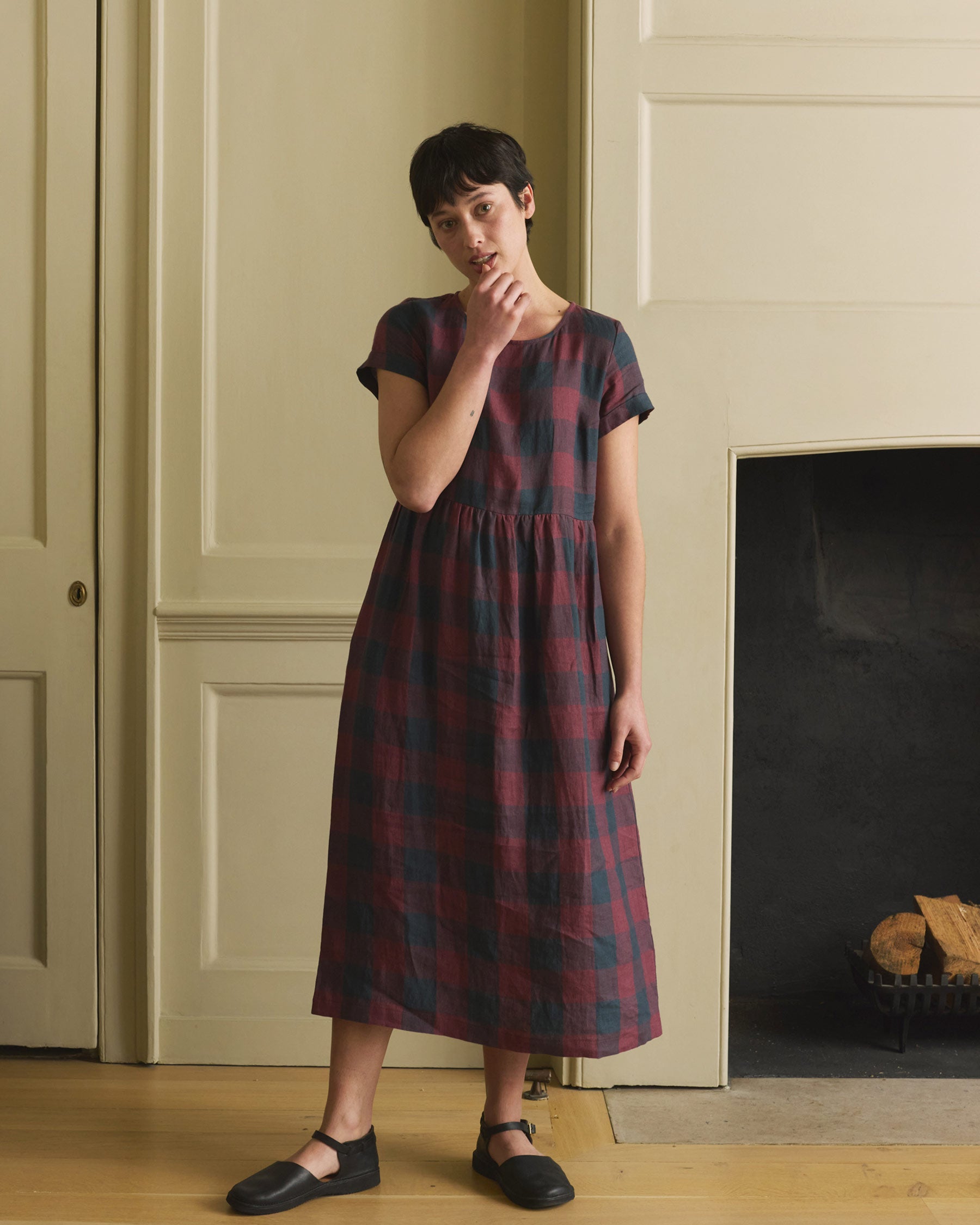 A person with short dark hair stands indoors by a fireplace, wearing the Pyne and Smith Clothiers No.17 Full Length Linen Tea Dress in Mulberry Check, vintage-inspired in dark red and navy, with one hand touching their chin.