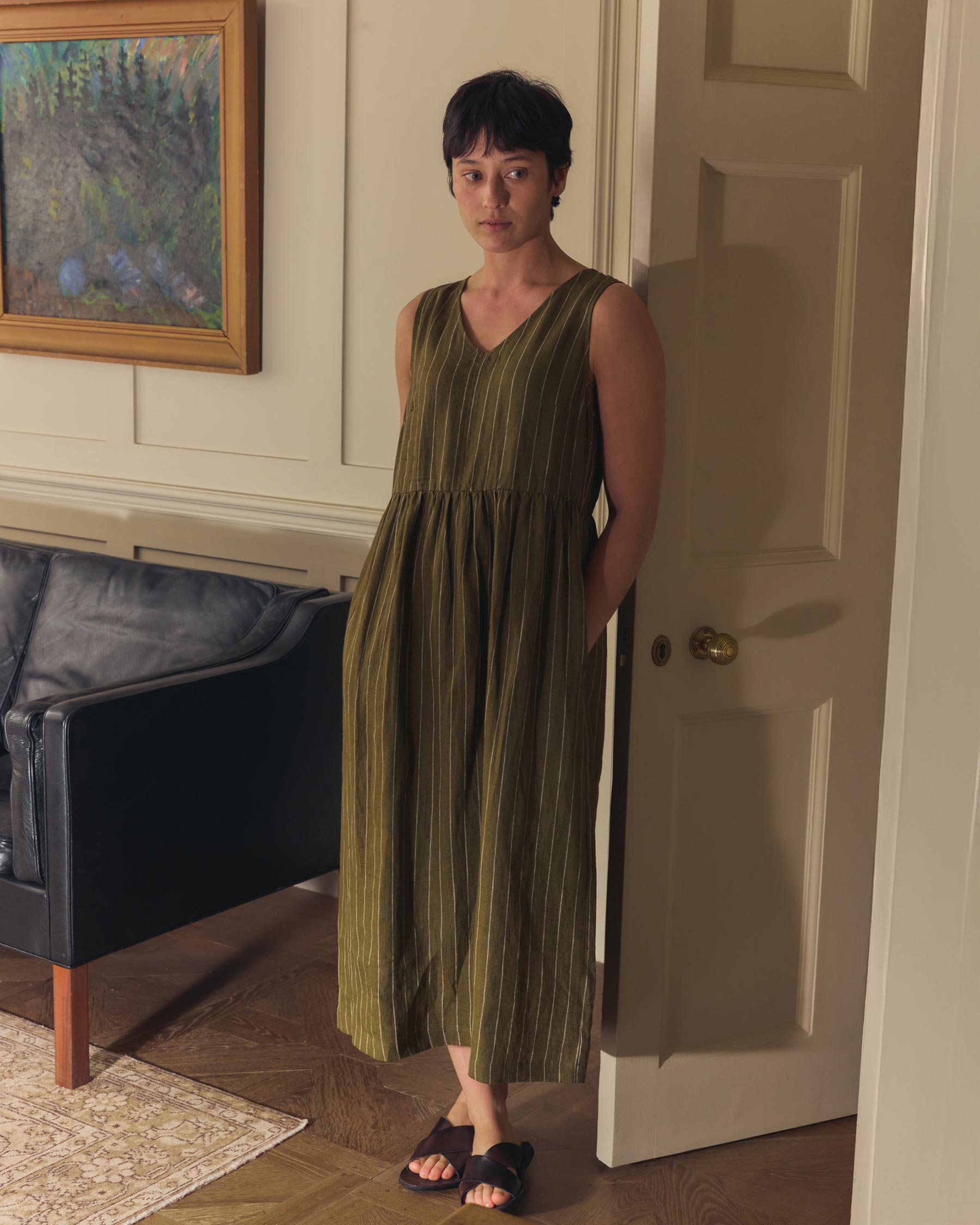 A person with short dark hair stands indoors by a door, wearing the No.21 V Neck Sleeveless Linen Dress in Kelp Green Stripe by Pyne and Smith Clothiers with black sandals. A dark sofa, framed artwork, and a rug are visible in the background.