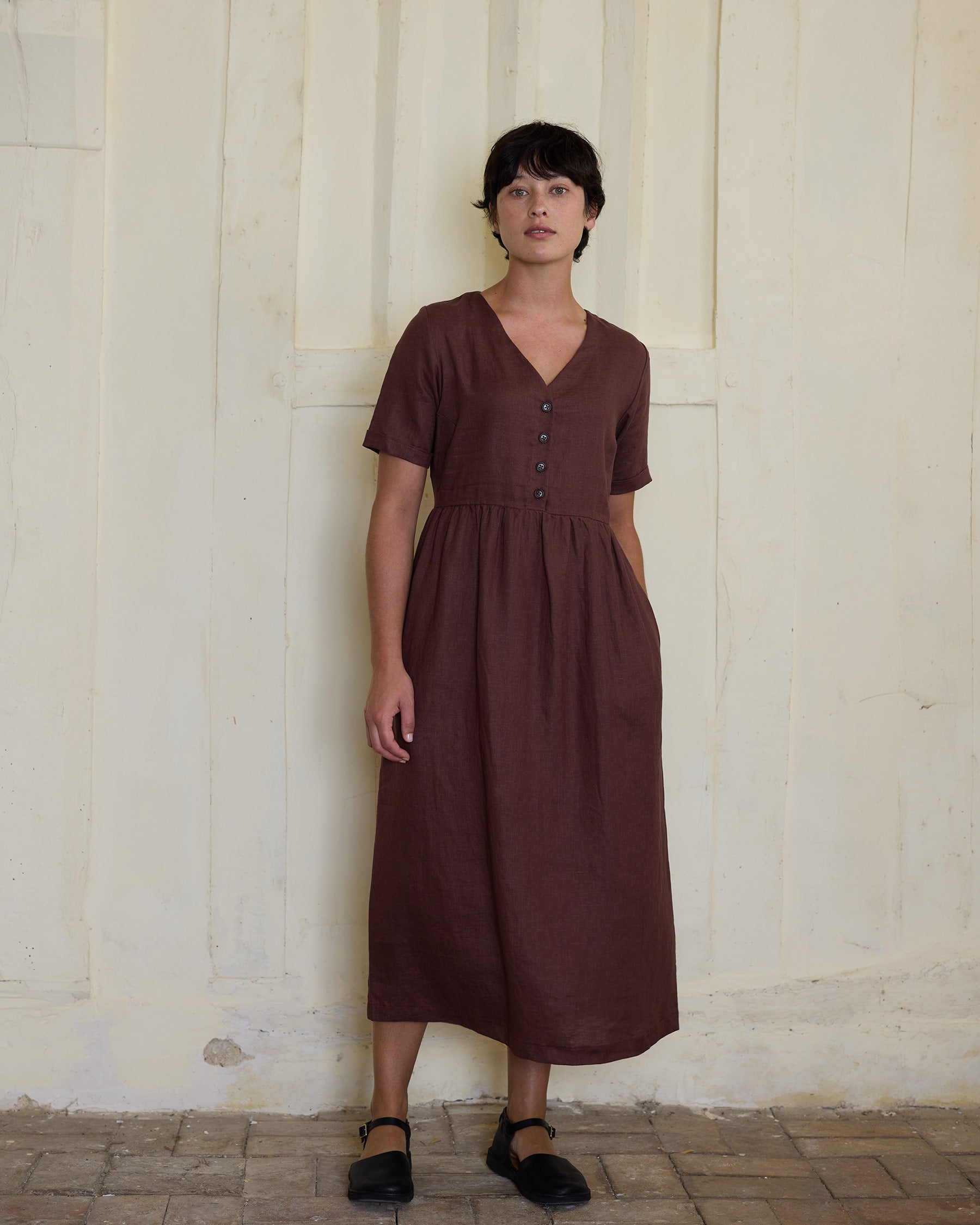 A person with short dark hair wears the Pyne and Smith Clothiers No.31 V Neck, Full Length Linen Dress in Marron and black shoes, standing against a textured light beige wall and looking at the camera.