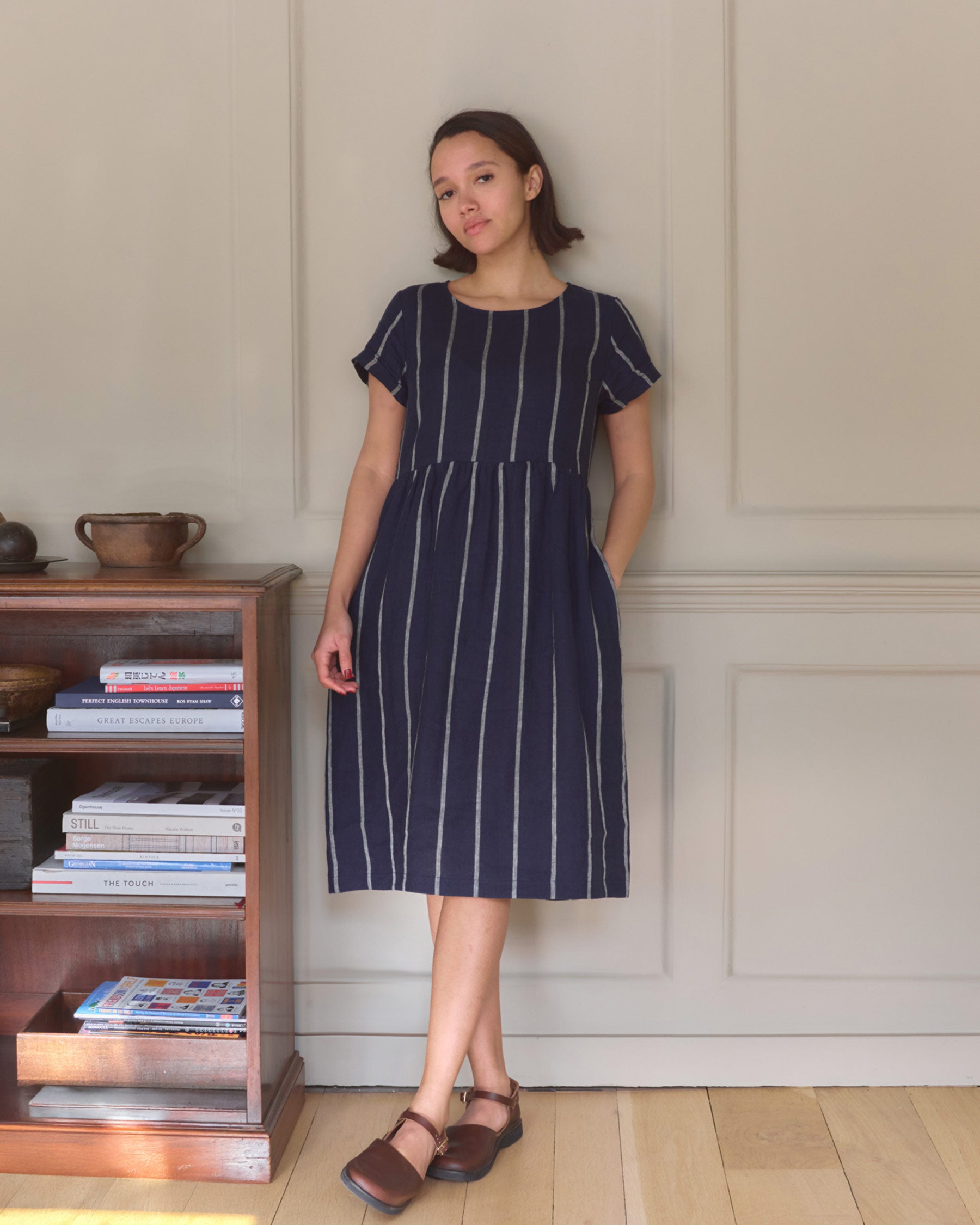 A woman stands indoors by a wooden bookshelf, relaxed in the Pyne and Smith Clothiers No.15 Linen Day Dress in Blundell Stripe—navy blue with white stripes, short-sleeved, handcrafted in California for timeless comfort.