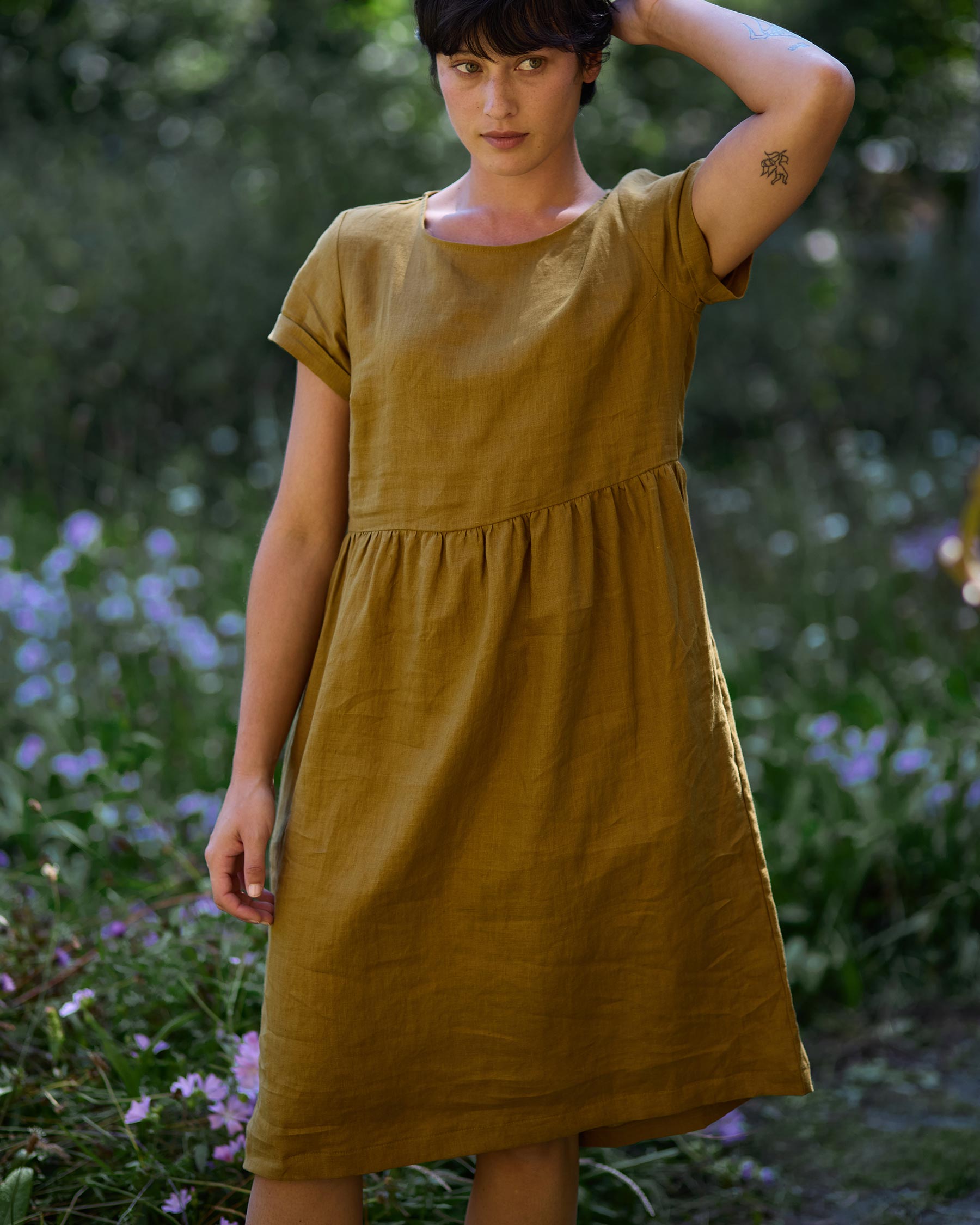 A person with short dark hair stands in a garden wearing the No.15 Linen Day Dress in Cumin by Pyne and Smith Clothiers, a sustainably made, loose-fitting piece. Purple flowers are visible in the background as they raise one hand to their head.