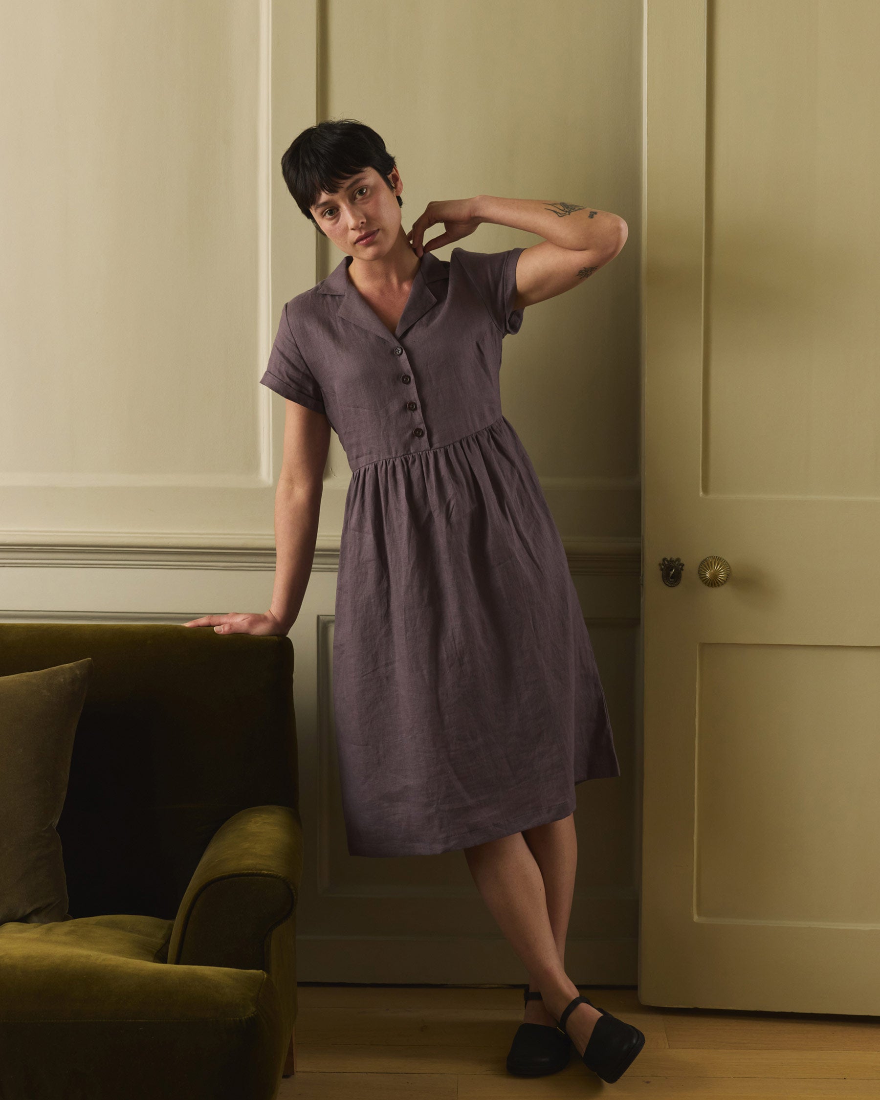 A person with short dark hair wears the Pyne and Smith Clothiers No.30 V-neck, Collared Linen Dress in Plum Truffle, standing indoors by a green sofa against a beige paneled wall, gently touching their neck.
