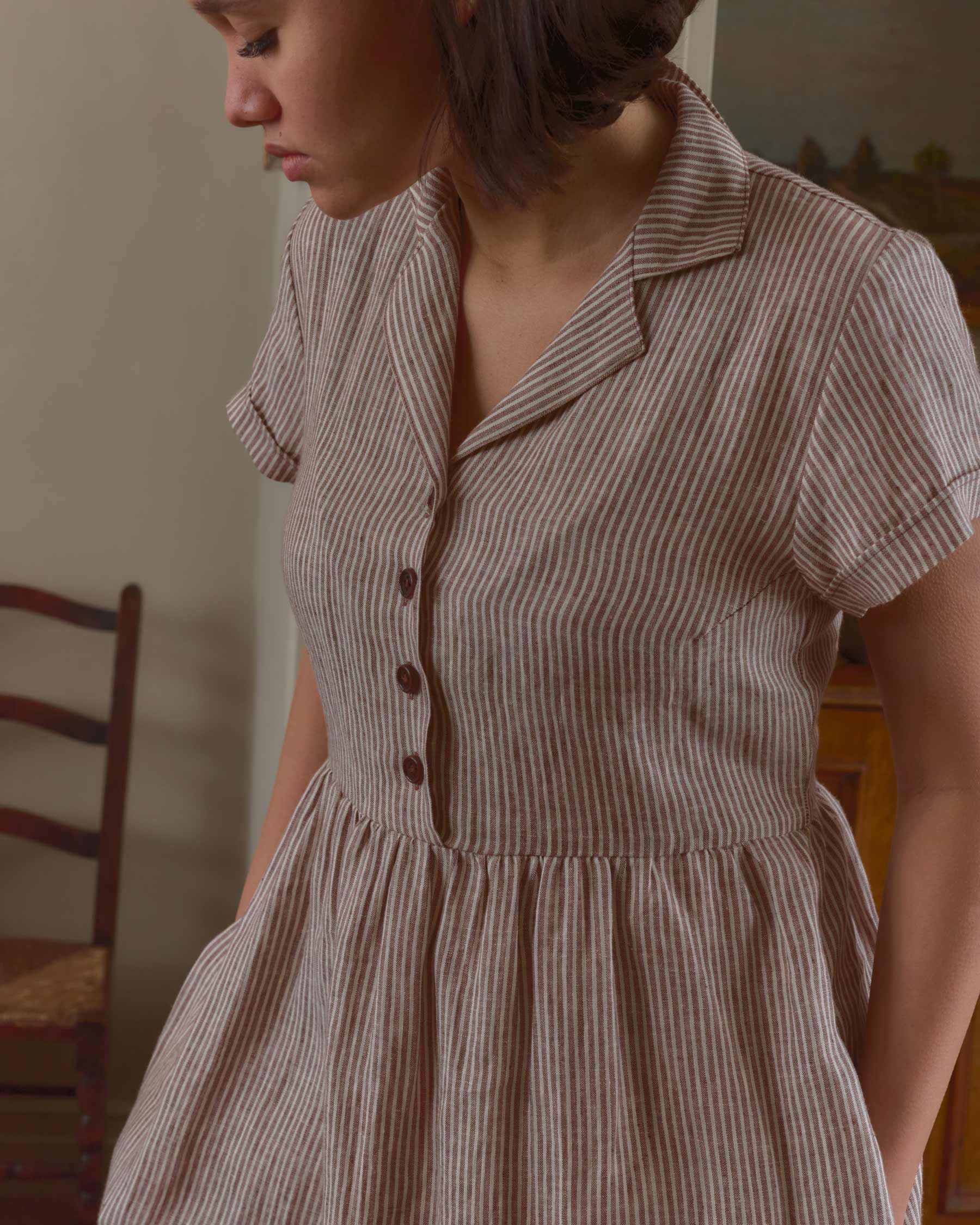 A woman stands indoors wearing the Pyne and Smith Clothiers No.30 Collared Linen Dress in Potters Stripe, a vintage-inspired short-sleeved brown and white striped shirt dress. A wooden chair and painting are in the background.