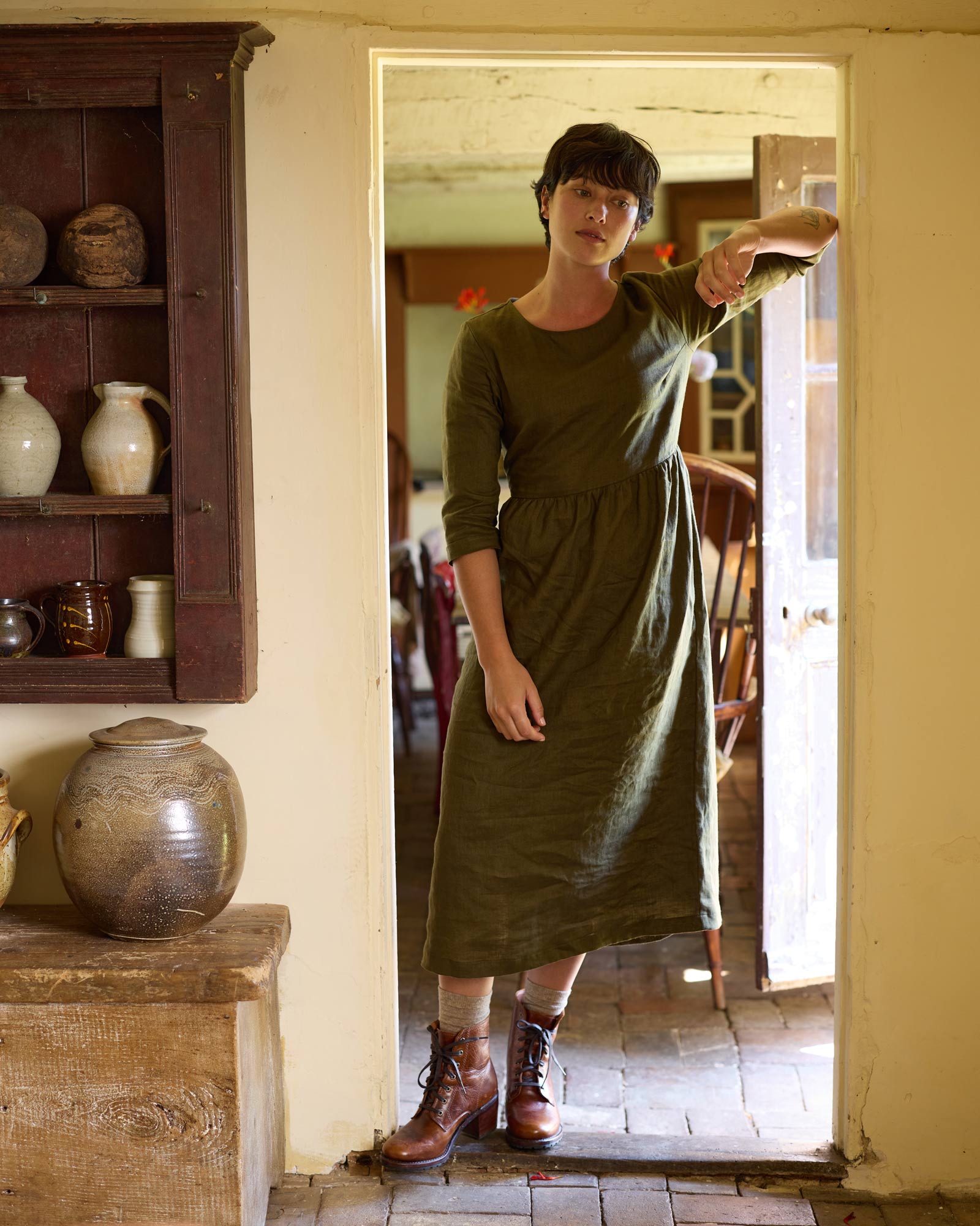 Wearing the No.41 Full Length Linen Day Dress in Beech by Pyne and Smith Clothiers, a person in brown boots stands in a rustic doorway, sunlight highlighting shelves of pottery and a large jug in the cozy room.