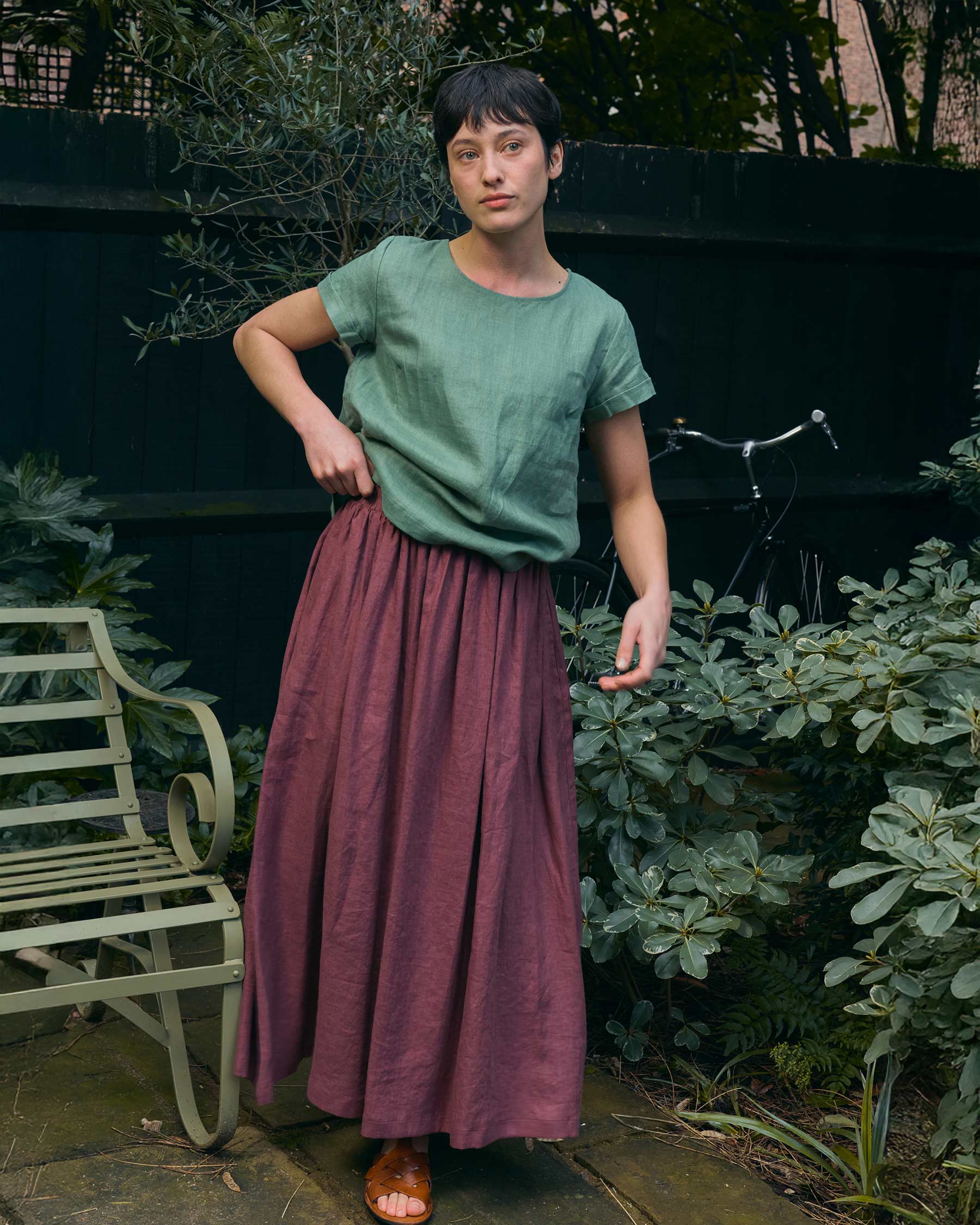 A person stands outdoors on a stone patio with green plants, wearing a green top, the Pyne and Smith Clothiers Linen Maxi Length Skirt No.12 in Wild Ginger, and brown sandals. A green bench and part of a bicycle are behind them.