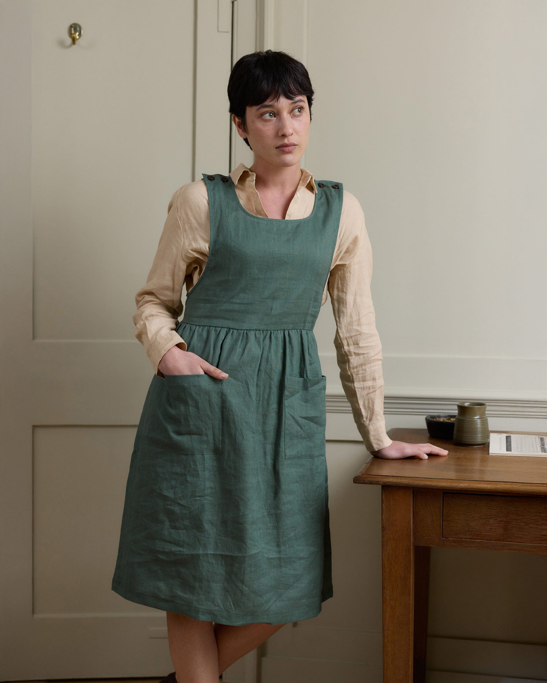 A person with short dark hair wearing a beige long-sleeve shirt and the Pyne and Smith Clothiers No.32 The Pinafore Dress in Carriage Green, made from sustainably grown European linen, stands by a wooden table in a softly lit room with pale walls.