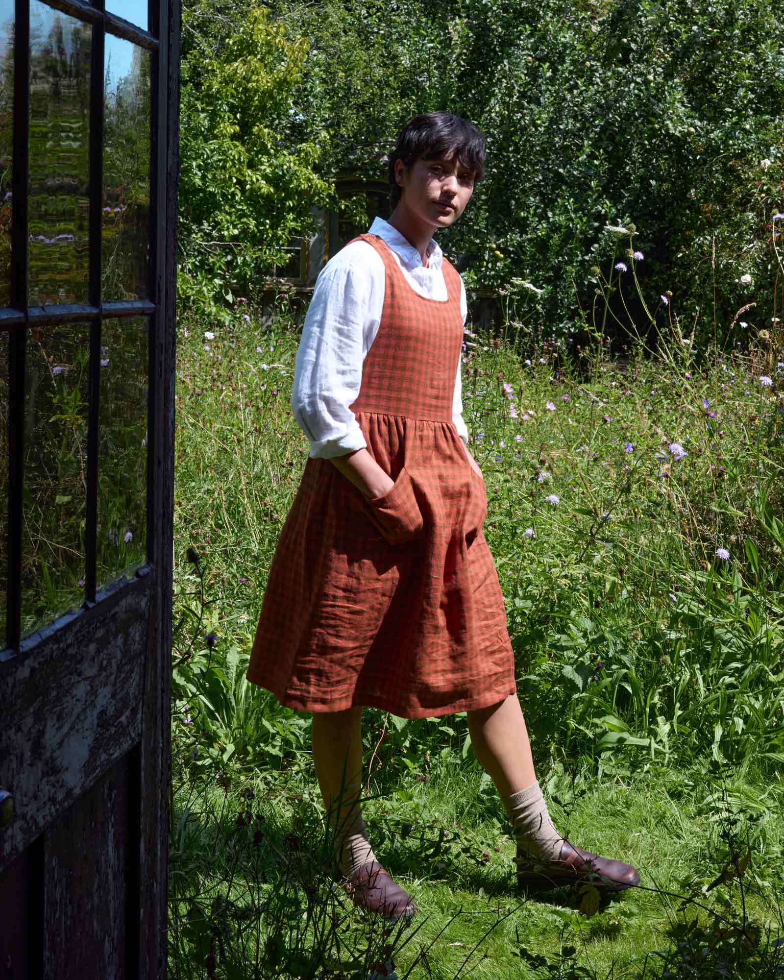 A person stands in a grassy garden, wearing the No.32 The Pinafore Dress in Ginger Check by Pyne and Smith Clothiers over a white shirt, with socks and brown shoes. Sunlight filters through trees; an old doorframe is visible on the left.