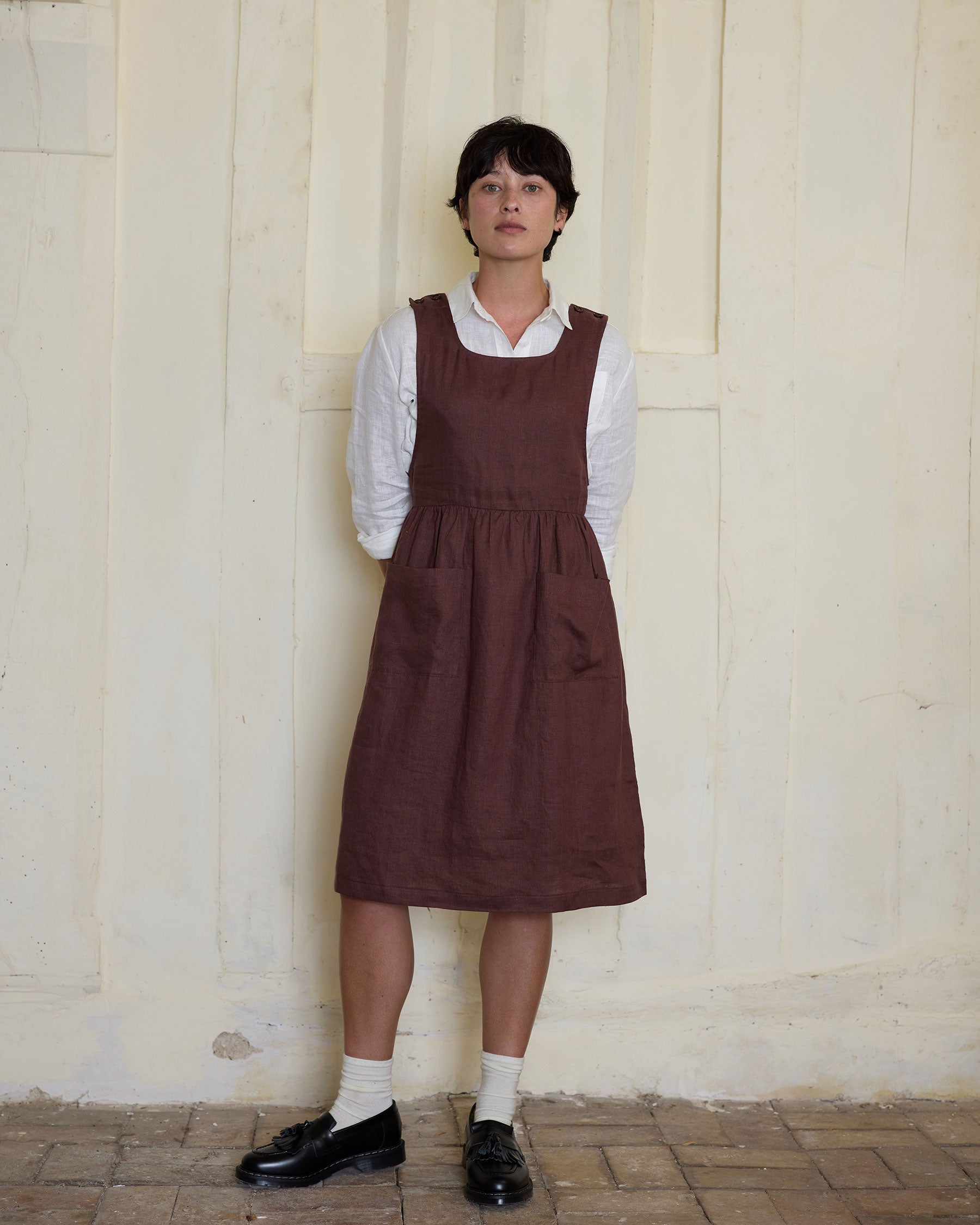 A person stands against a cream-colored textured wall, wearing Pyne and Smith Clothiers No.32 The Pinafore Dress in Marron over a white long-sleeve shirt, paired with white socks and black shoes, hands behind their back.