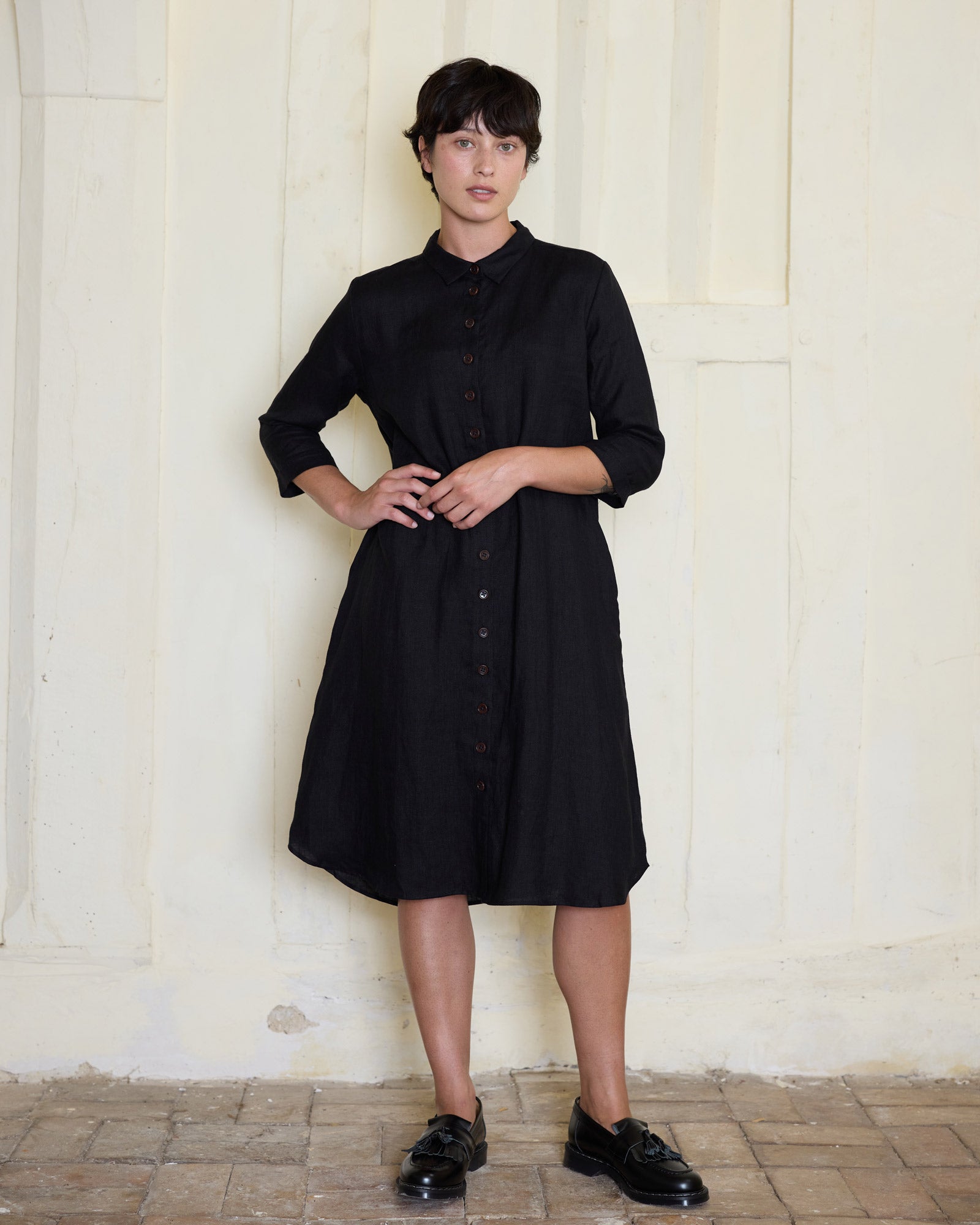 A person with short dark hair wears Pyne and Smith Clothiers’ No.39 The Linen Shirtdress in Black and matching shoes, standing against a pale textured wall; the button-up is made from sustainably grown linen.