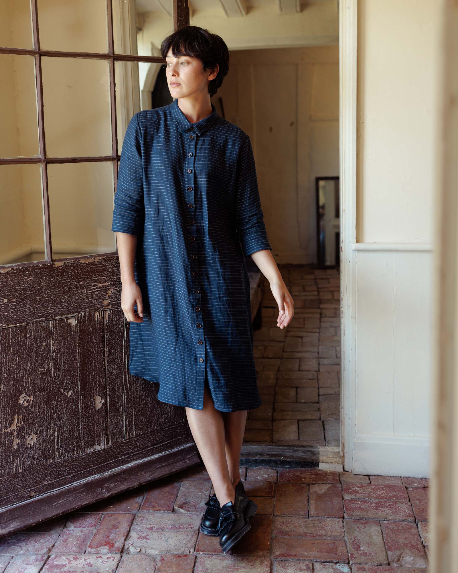 A person in Pyne and Smith Clothiers No.39 The Linen Shirtdress in The Swedish Blue, paired with black shoes, stands indoors on a brick floor against a weathered door, relaxed, highlighting the dress’s subtle embroidered stitch detailing.