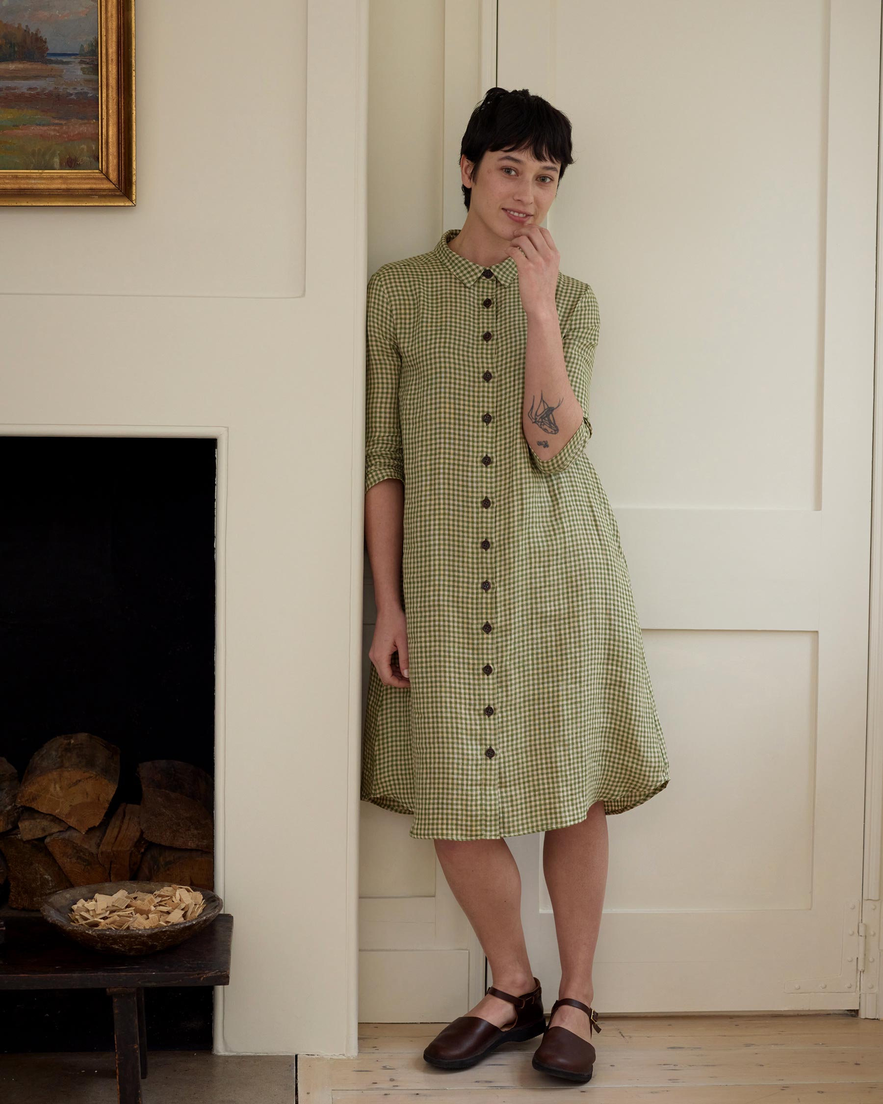 A woman with short dark hair stands indoors against a cream wall, wearing the Pyne and Smith Clothiers No.39 The Linen Shirtdress in Wimbledon with 3/4 sleeves and brown shoes. She touches her face and smiles; logs are stacked in the open fireplace nearby.