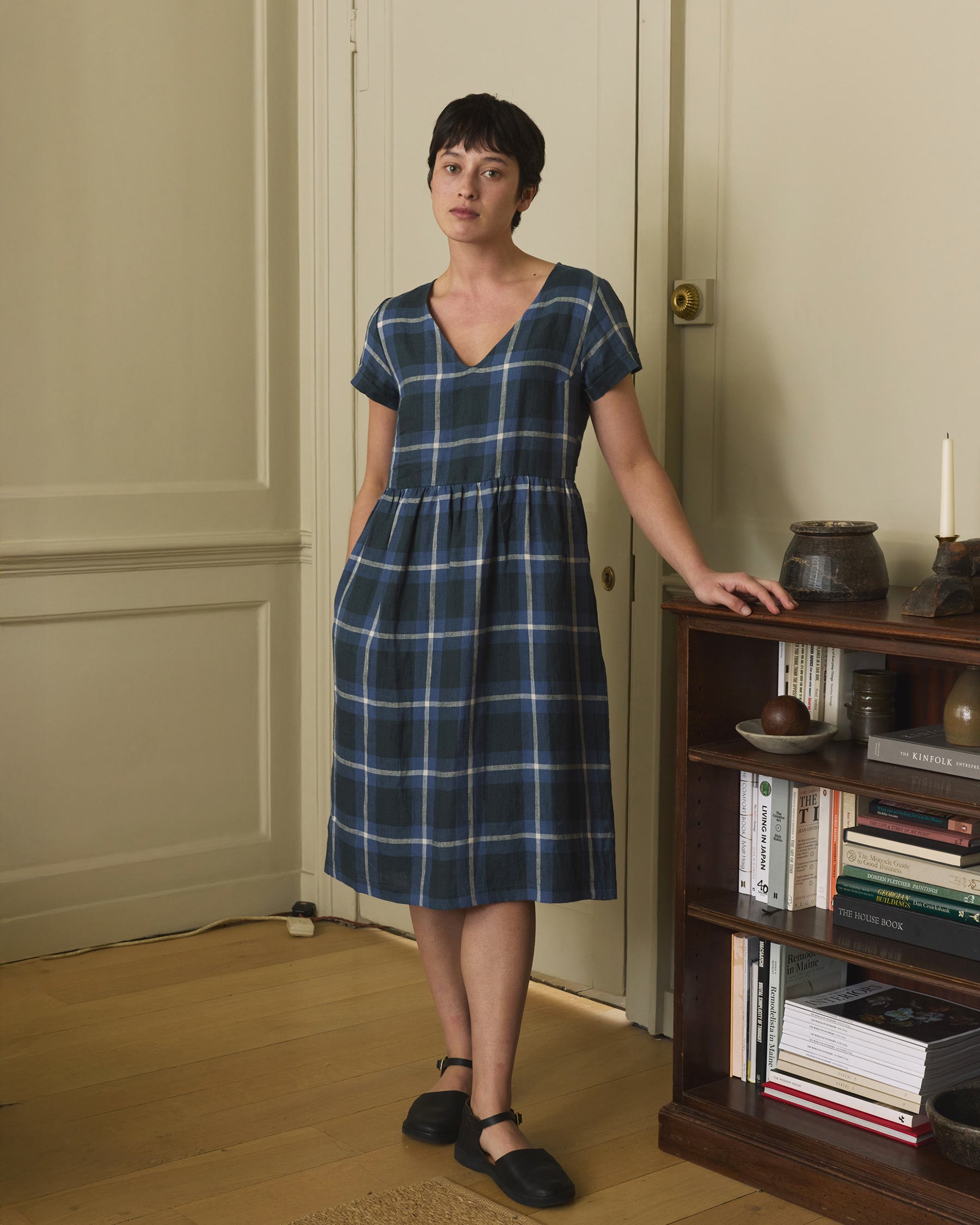 A person with short dark hair, wearing the Pyne and Smith Clothiers No.40 V Neckline Linen Day Dress in Stirling Plaid and black shoes, stands indoors by a wooden bookshelf filled with books and pottery in a calm, homey setting.