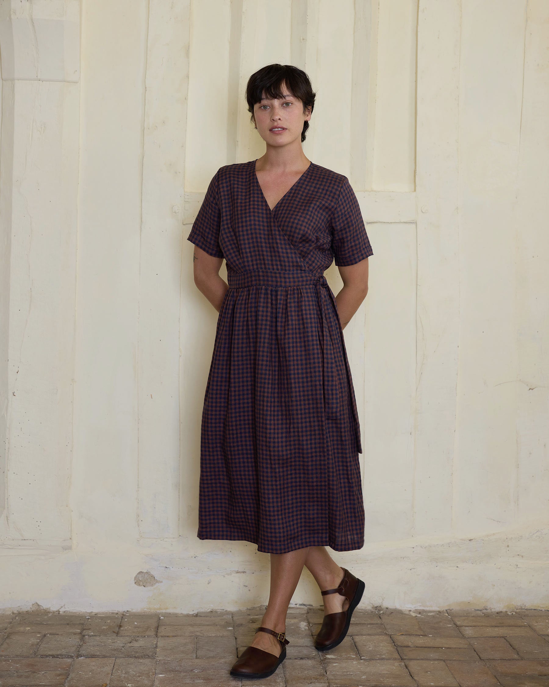A person with short dark hair stands against a pale, textured wall wearing the Pyne and Smith Clothiers No.37 Wrap Style Linen Dress in Princelet Check, paired with brown strap shoes and hands behind their back.