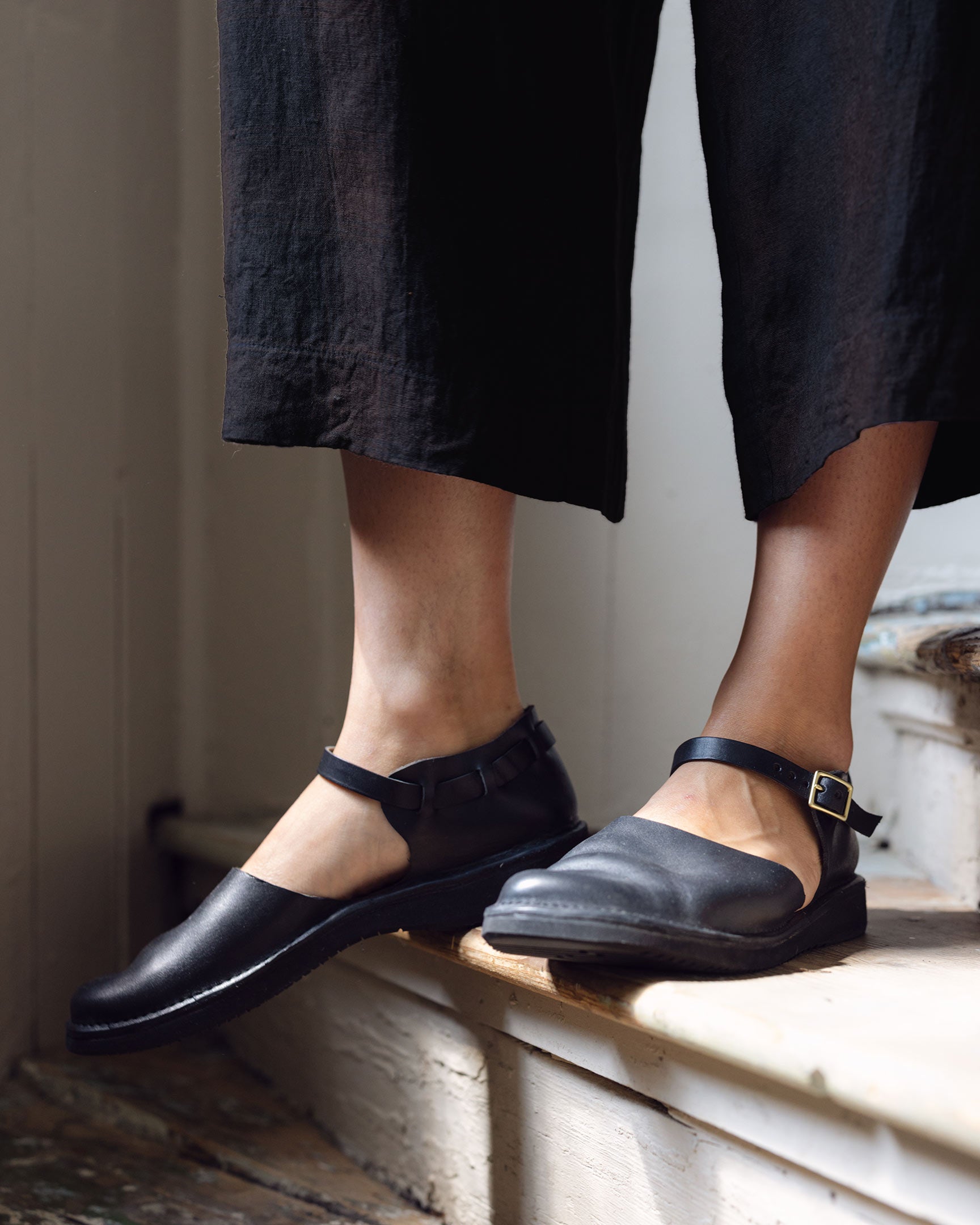 A person wearing black, wide-leg pants and black Mary Jane shoes stands on wooden steps indoors, one foot on a higher step and the other on a lower step.