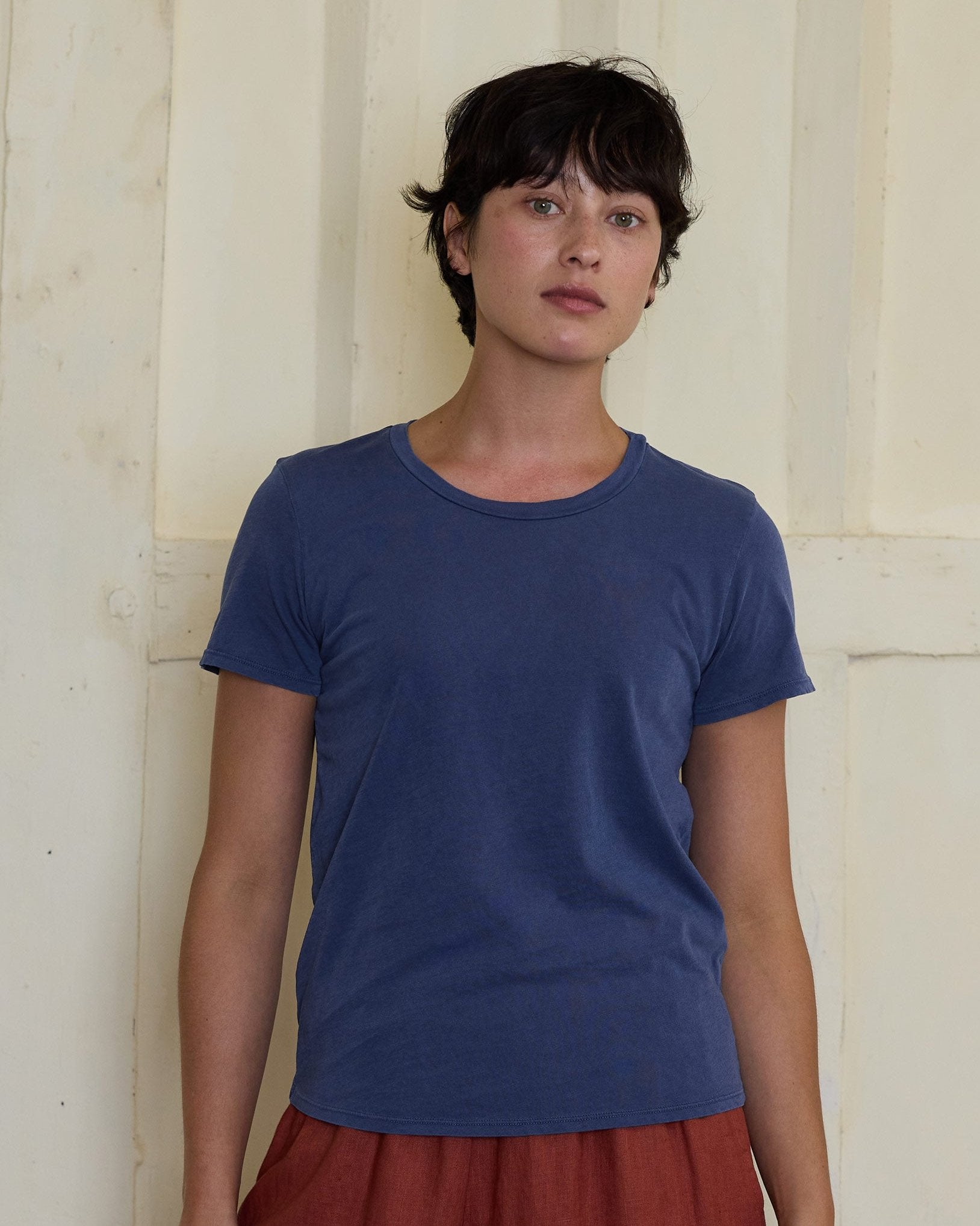 A person with short dark hair wears a Pyne and Smith Clothiers Organic Cotton T-shirt in Vintage Wash Peony Blue and rust-colored pants, standing before a light textured wall, looking at the camera with a casual vintage style.