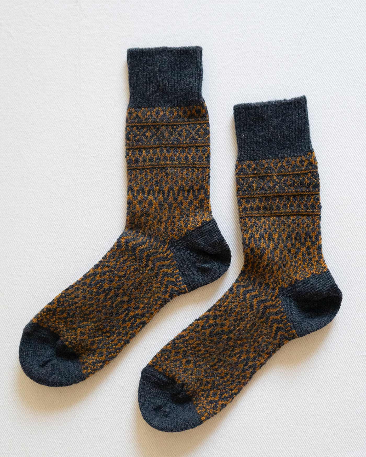 A pair of Pyne and Smith Clothiers Wool Jacquard Socks in Navy, featuring mid-calf length with cozy jacquard knit, navy base, and a brown geometric pattern with stripes and zigzags, laid flat on a light background.
