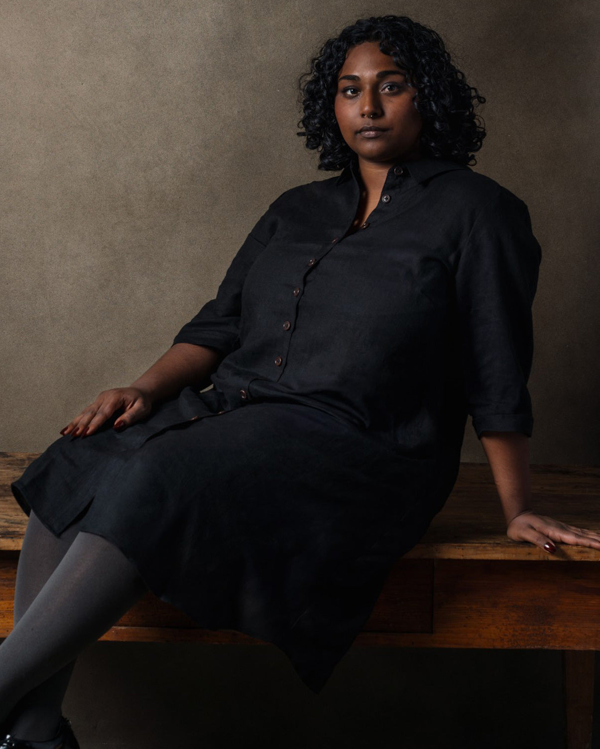 A person with curly dark hair sits on a wooden table against a plain brown background, confidently wearing the No.39 The Linen Shirtdress in Black by Pyne and Smith Clothiers.
