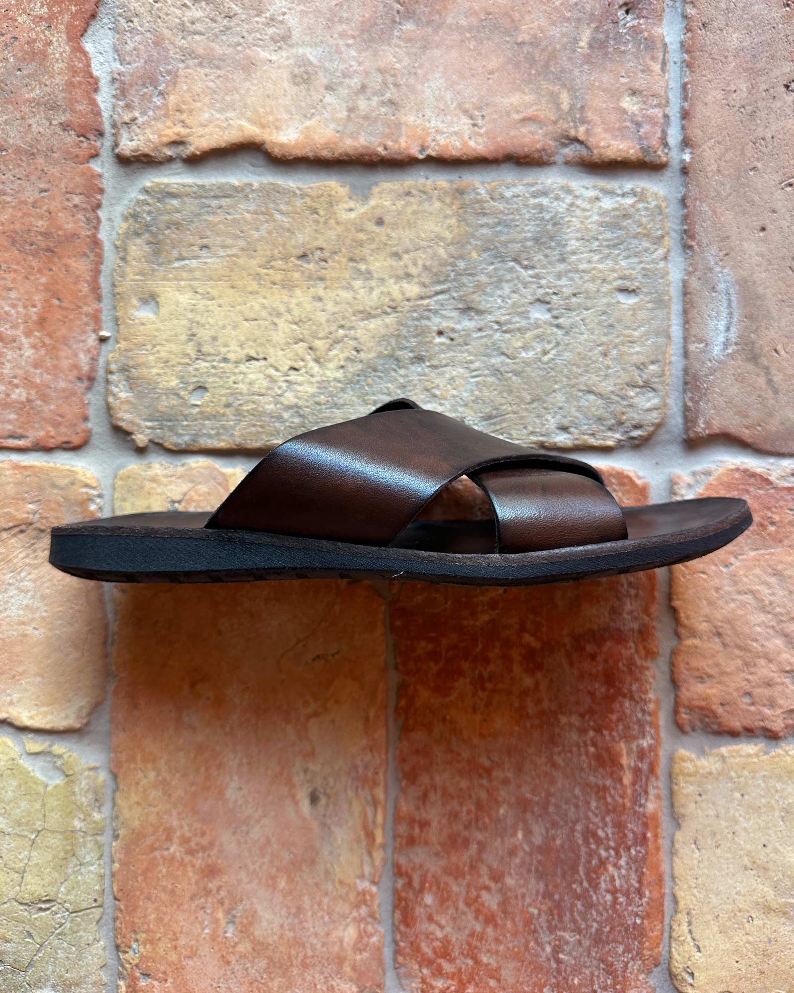 The Pyne and Smith Clothiers Dark Brown Leather Infinity Sandal, featuring crisscross leather straps and a premium upper, is shown sideways against a wall of multicolored textured stone tiles.